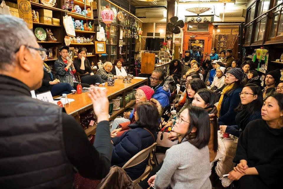 A man is speaking to a large audience in a cozy room decorated with shelves of collectibles and Asian art. The audience includes diverse individuals sitting and listening attentively.