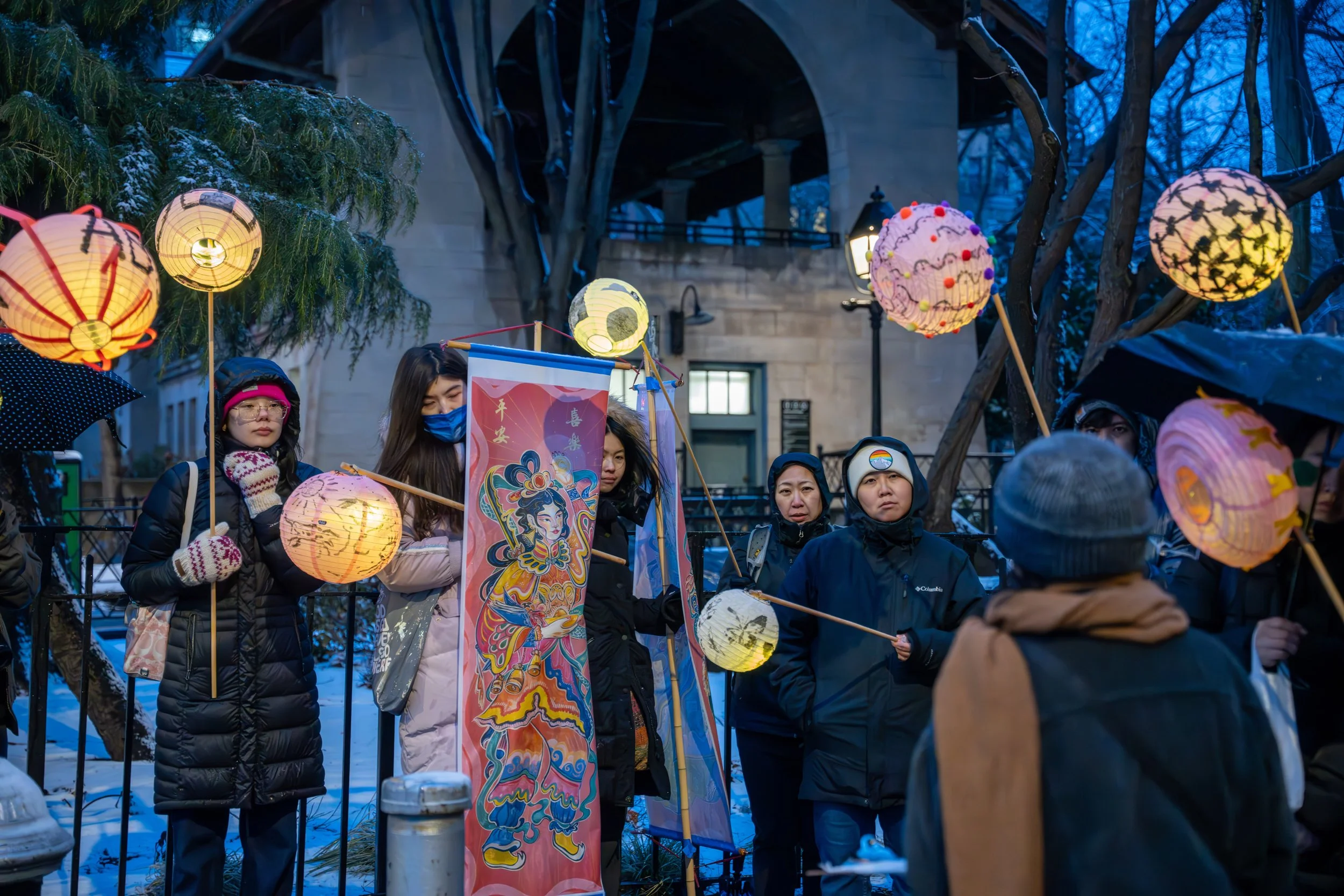 People gathered outdoors at dusk, holding colorful paper lanterns for a celebration, with a banner featuring traditional artwork and snow on the ground.