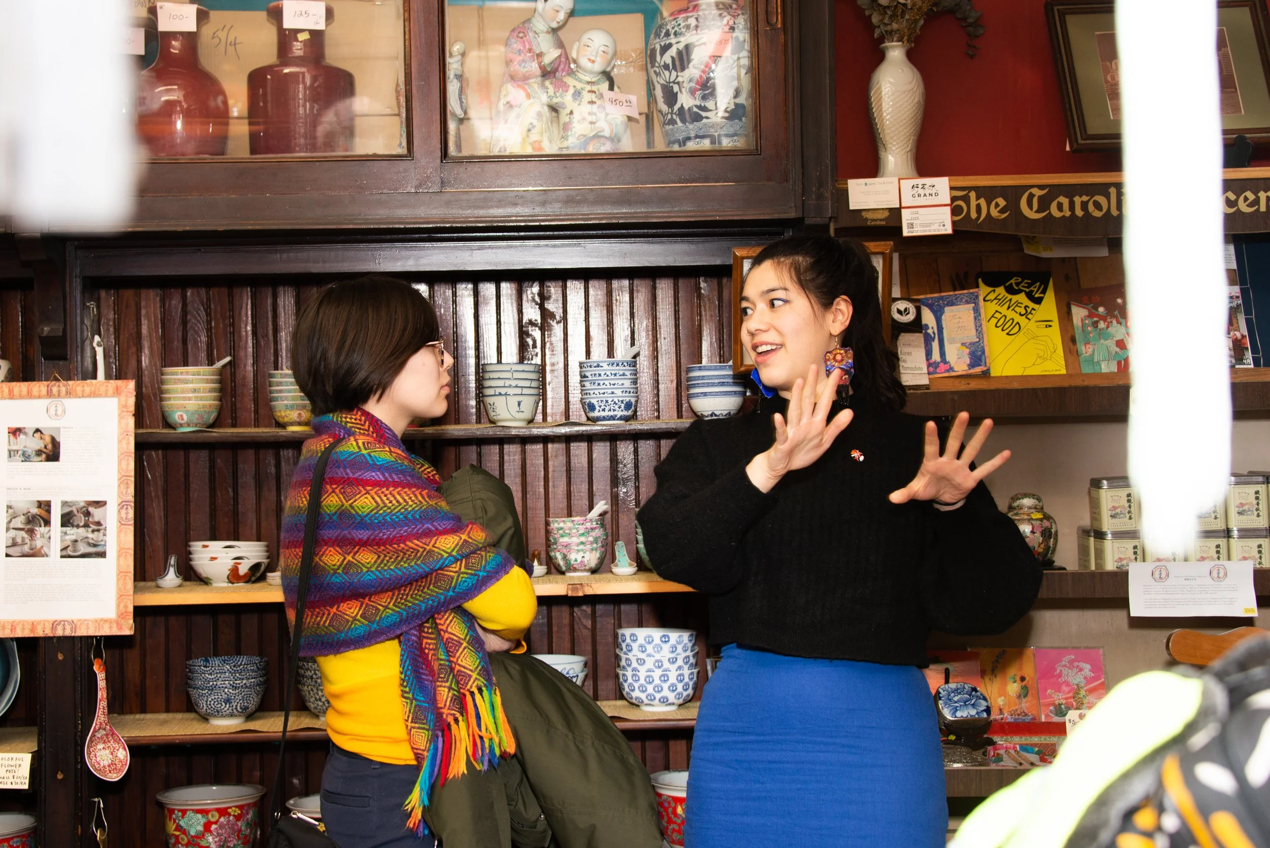 Two women are inside a store with shelves filled with Chinese and Japanese ceramics. The woman on the right is talking and gesturing with her hands, wearing a black sweater and a blue skirt. The woman on the left is listening, wearing glasses, a colorful shawl, and a yellow top.