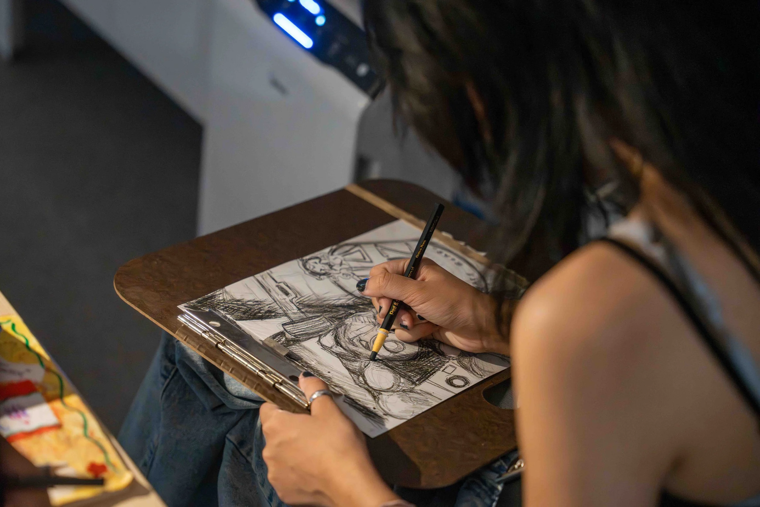 A person drawing comic book characters on a sketchpad using a black marker. The person is sitting at a wooden table, and the drawing includes a shirtless male figure and a female character. The person has dark hair and is wearing a black sleeveless top.