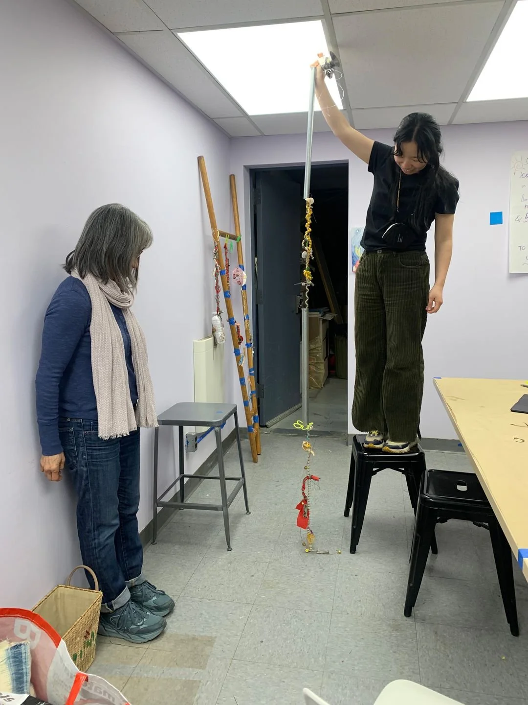 A young woman standing on a chair holding a long string of objects hanging from the ceiling, while an older woman observes in a room with various items and decorations.