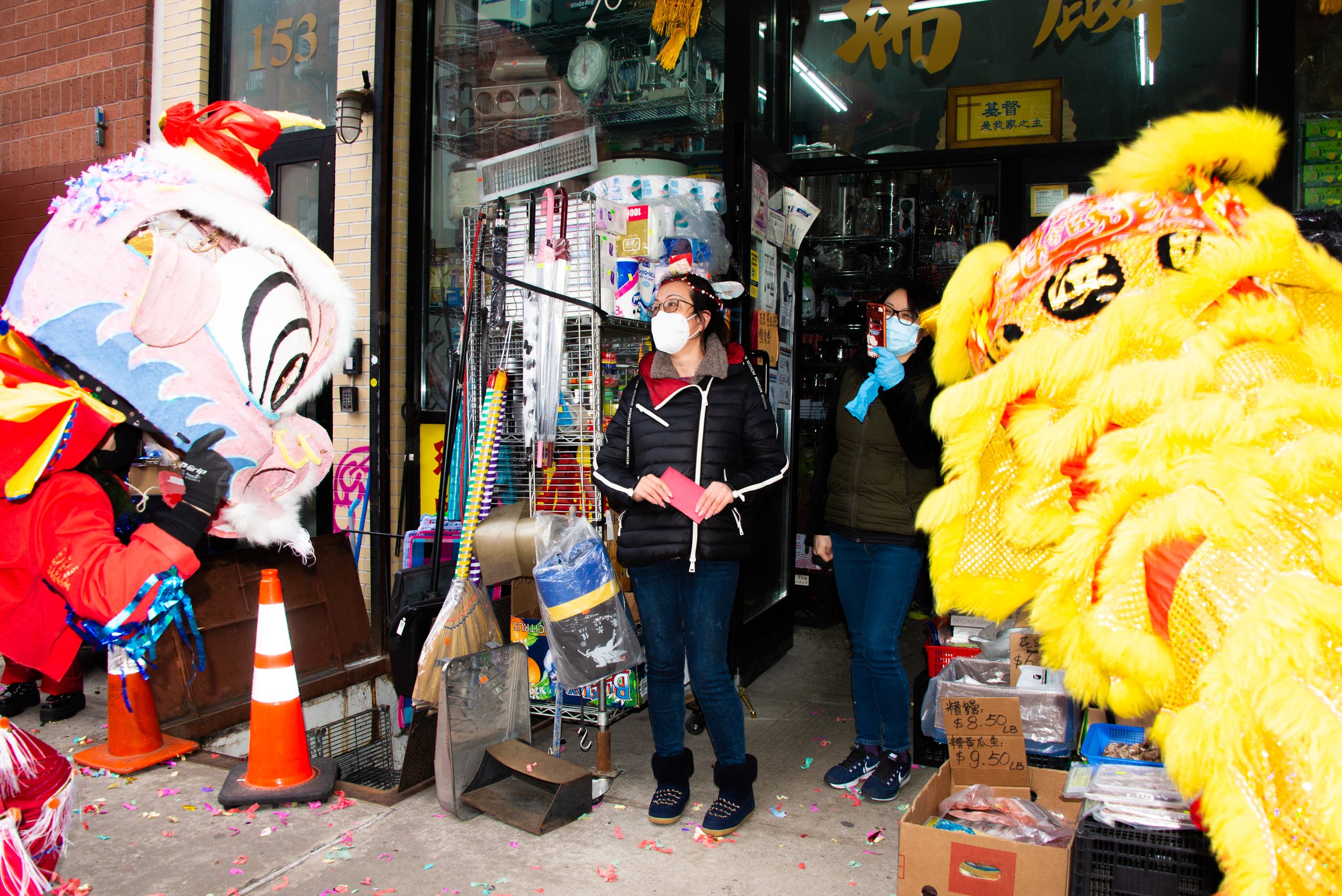People celebrating a Chinese New Year festival outside a store, with two individuals in traditional lion dance costumes, one pink and the other yellow, and two women wearing masks and winter clothing.