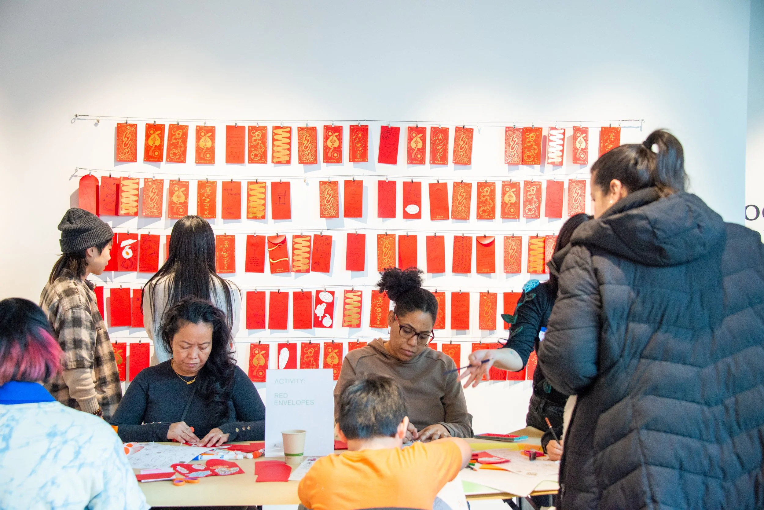 People participating in an activity at a table, with red envelopes displayed on a white wall behind them.
