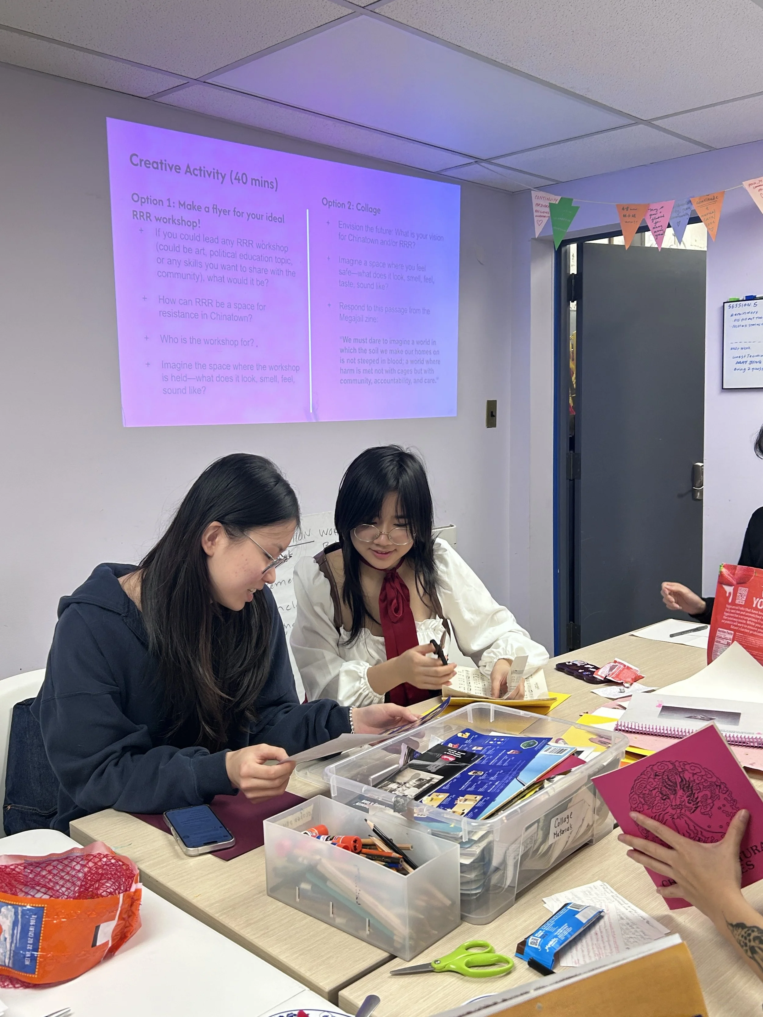 Two women sitting at a table working on creative activity, surrounded by supplies, with a screen showing workshop instructions in the background.