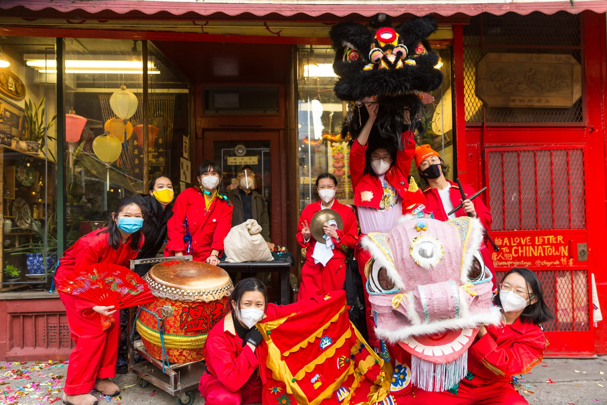 A group of people wearing red outfits celebrating a cultural event in front of a building decorated with hanging lanterns and traditional decor. Some are holding musical instruments, and a performer is dressed in a lion dance costume with a large pink and white lion head. Confetti is on the ground, and everyone is wearing face masks.