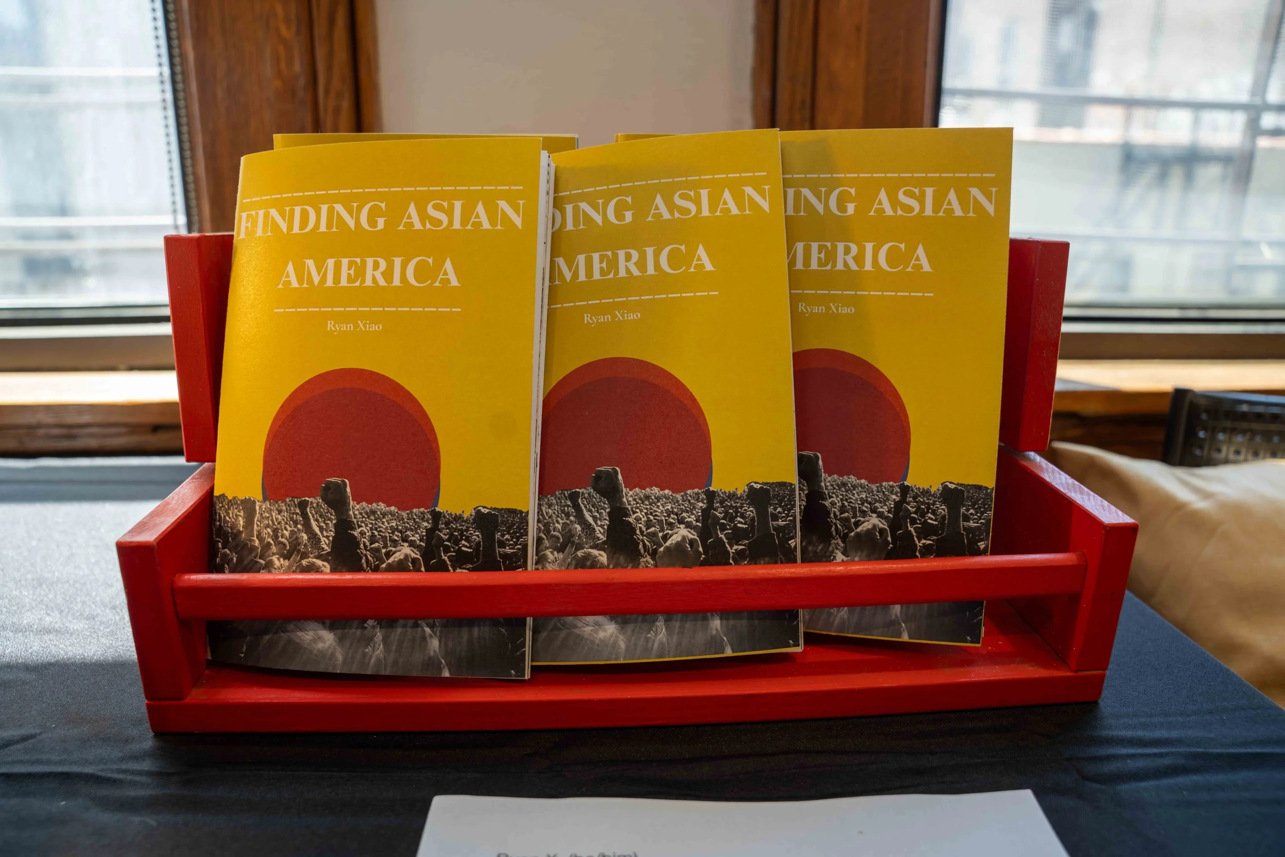 Books in a red display rack titled 'Finding Asian America' by Ryan Xiao, with a large yellow cover featuring an illustration of a large red circle and black and white photographs of protests.