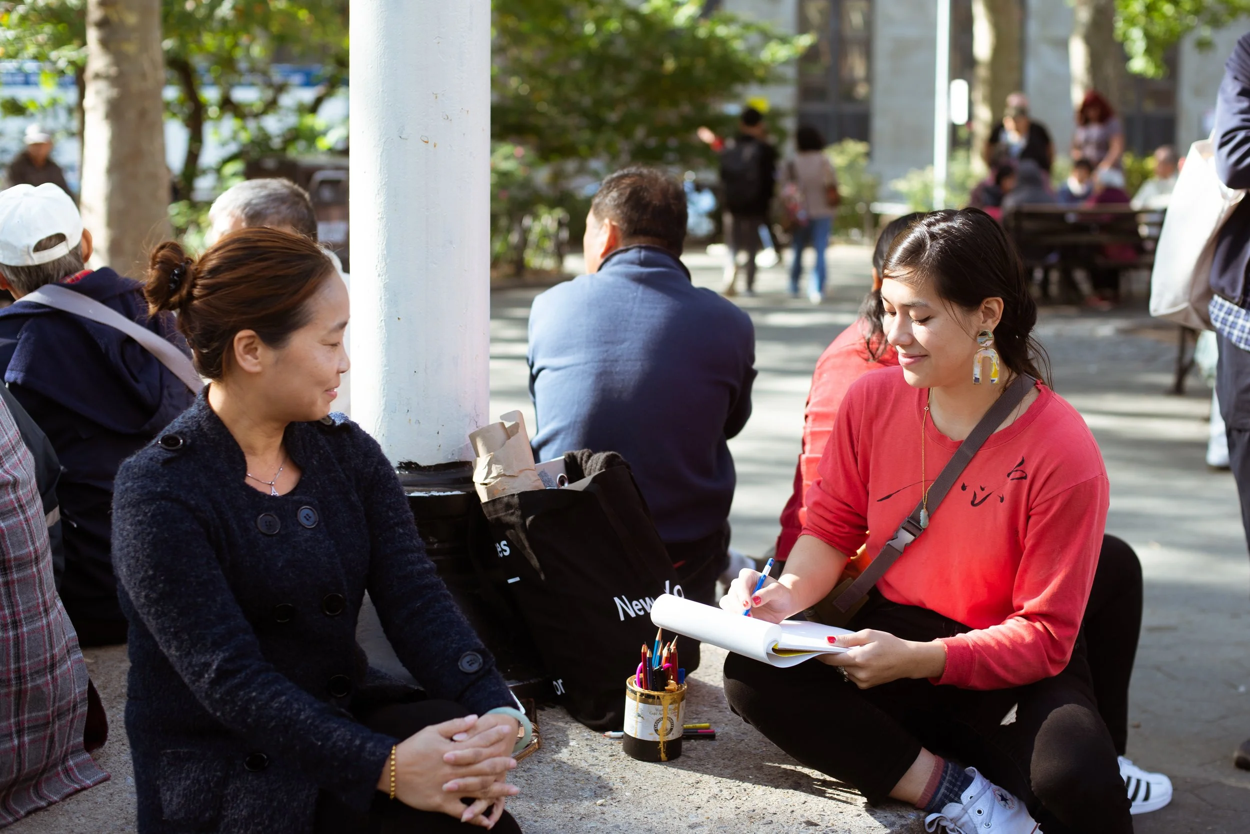 Two women sitting on the ground in a park, one appears to be conversing with the other who is taking notes in a notebook. There are people in the background walking and sitting on benches.