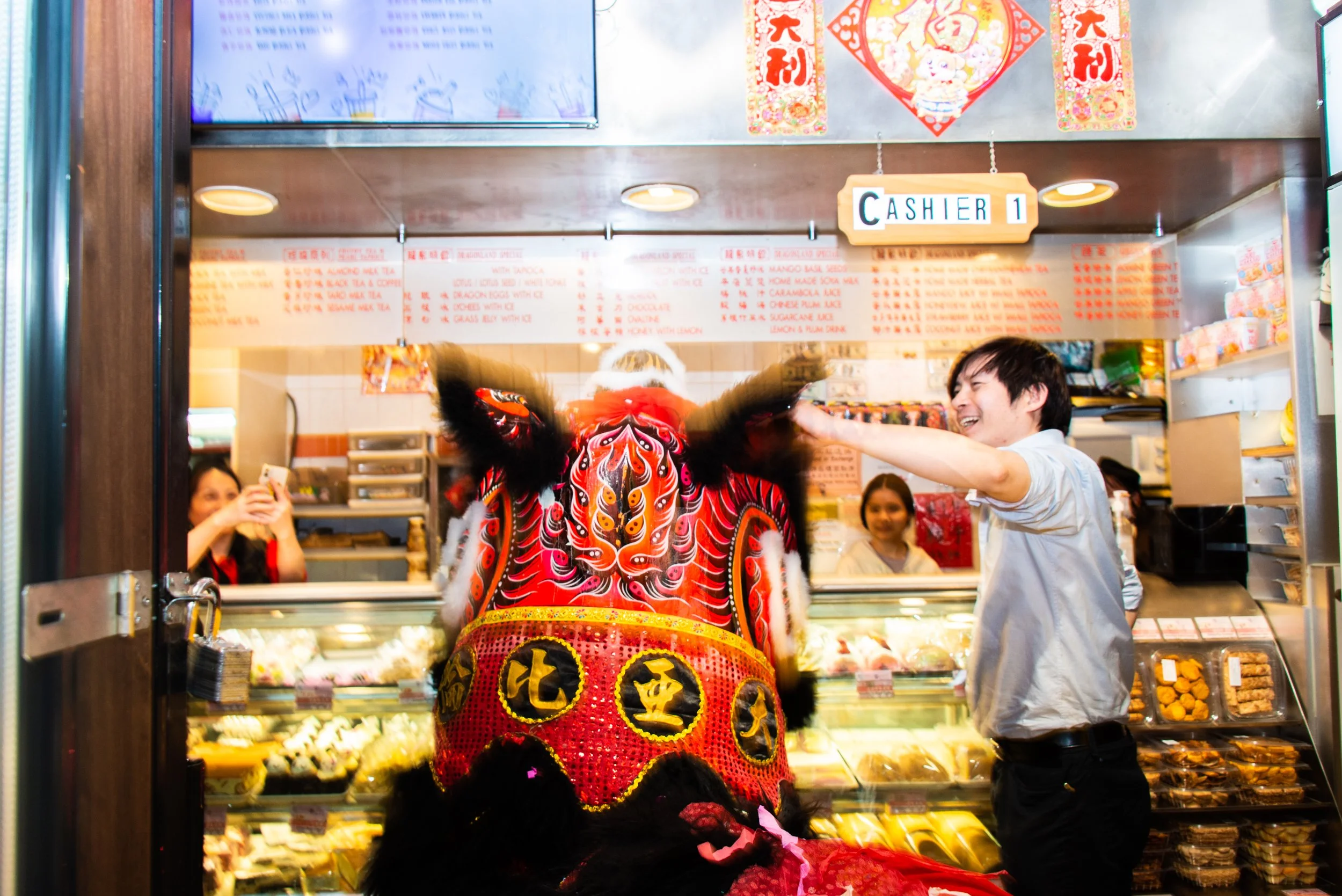 A person in a colorful lion dance costume inside a bakery, with a man smiling and reaching out to the costume. Several women are in the background, one taking a photo. The bakery has a sign labeled 'Cashier 1' and a menu board overhead.