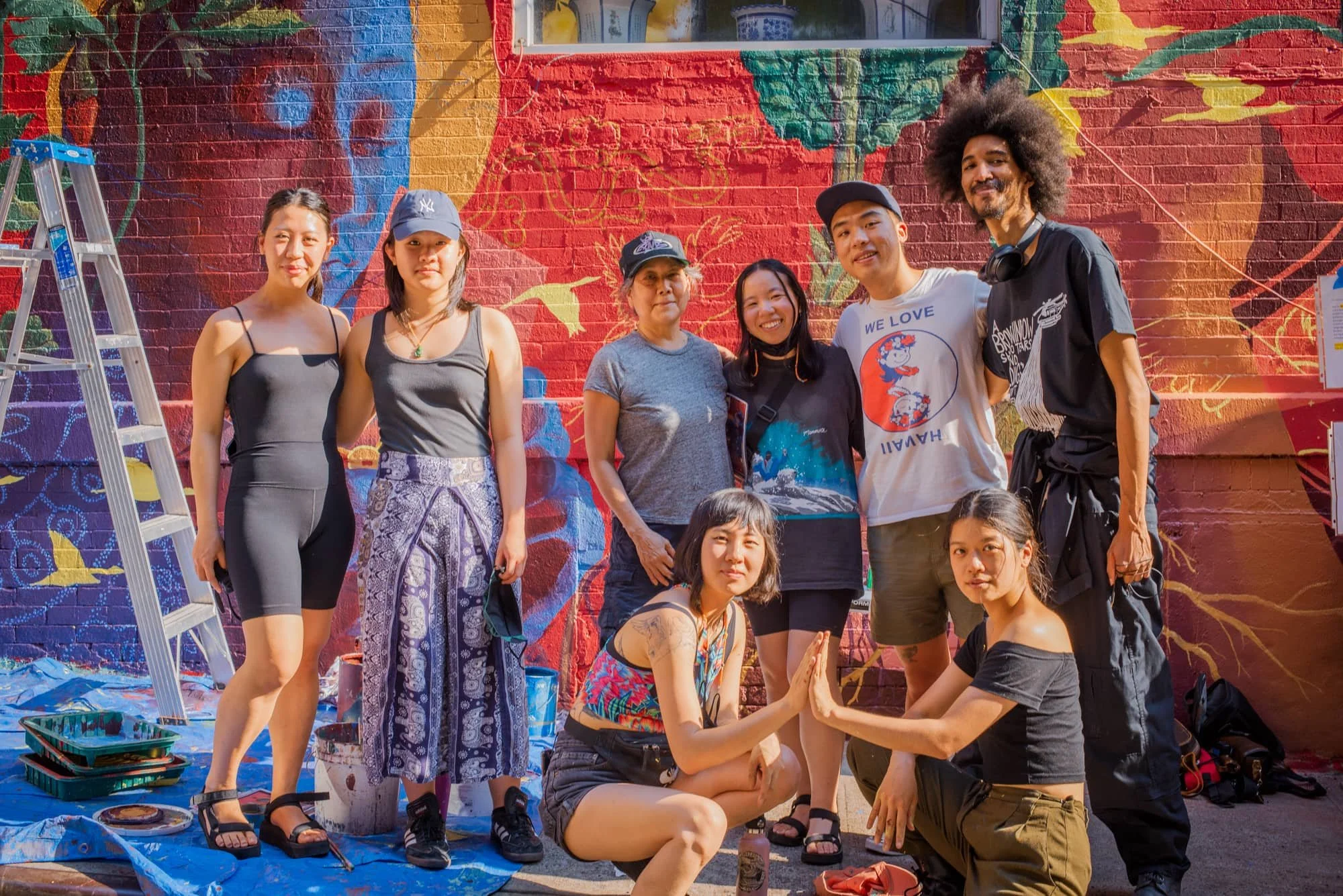 Group of diverse people painting a colorful mural on a brick wall outdoors. Some people are standing, while others are kneeling or sitting, and there are painting supplies on the ground.