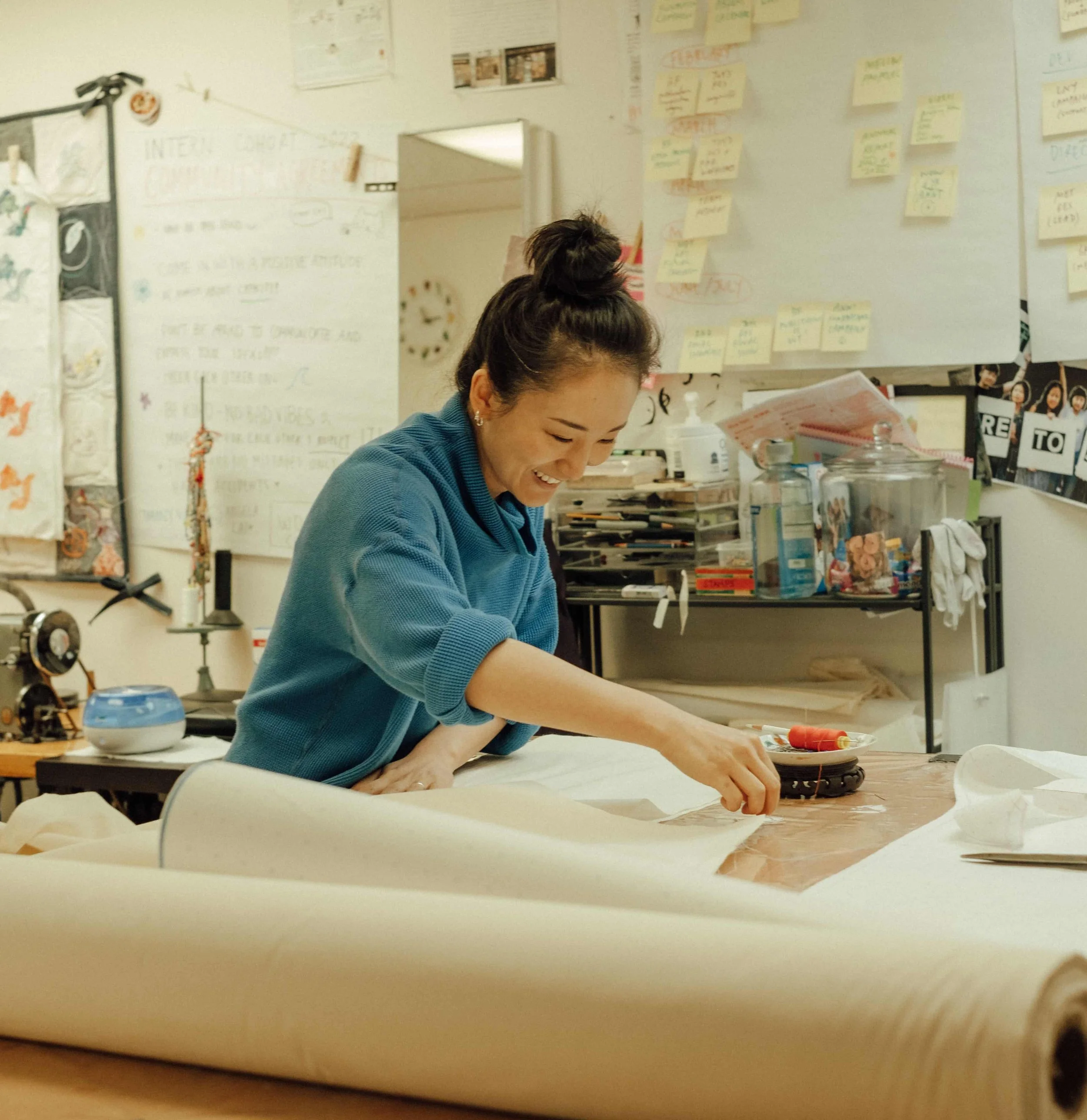 A woman in a blue sweater smiling and working at a large table covered with fabric in a crafts or design studio.