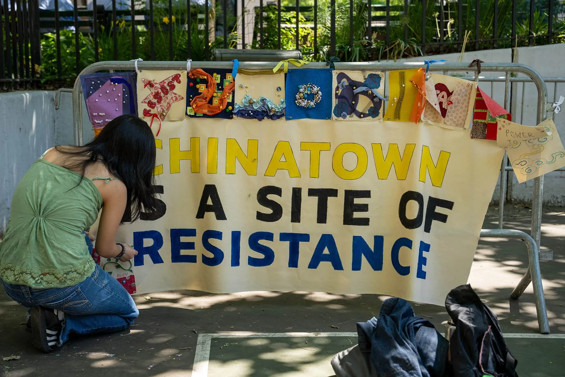 A young woman kneeling next to a large yellow banner that reads "Chinatown is a site of resistance" in bright yellow, black, and blue letters. The banner is decorated with colorful handmade crafts hanging along the top, including fabric art and jewelry. There are backpacks and bags on the ground nearby.