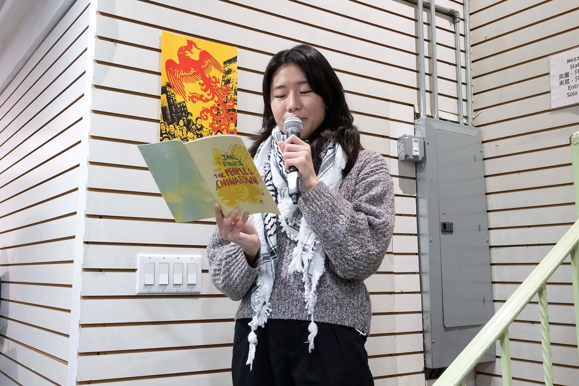 A woman with dark hair, wearing a gray sweater and a patterned scarf, is speaking into a microphone while holding a yellow booklet titled 'The People's Chinatown.' She stands in front of wood-paneled walls with a red and yellow poster behind her.