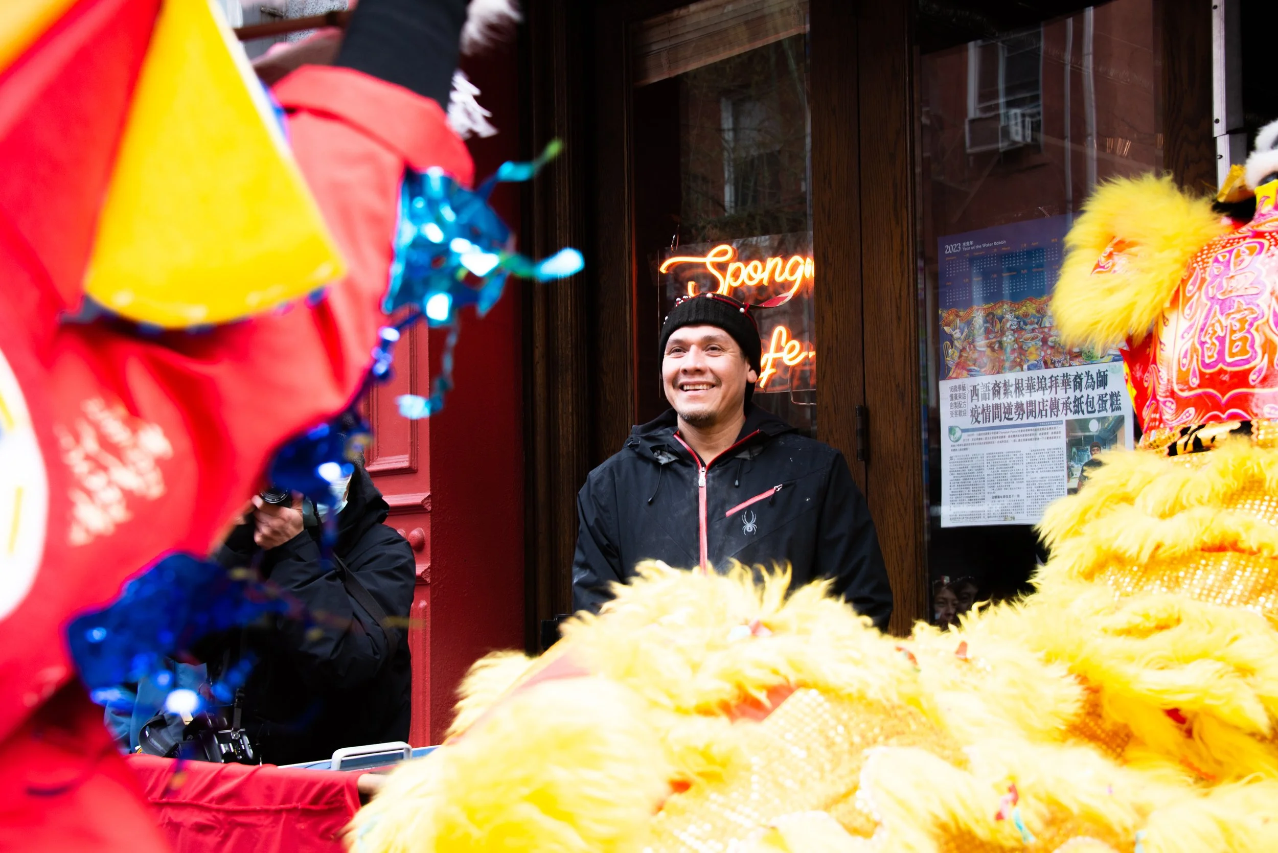 Smiling man in black jacket and black beanie standing outside a restaurant with Chinese signage, surrounded by people and bright yellow lion dance costumes.