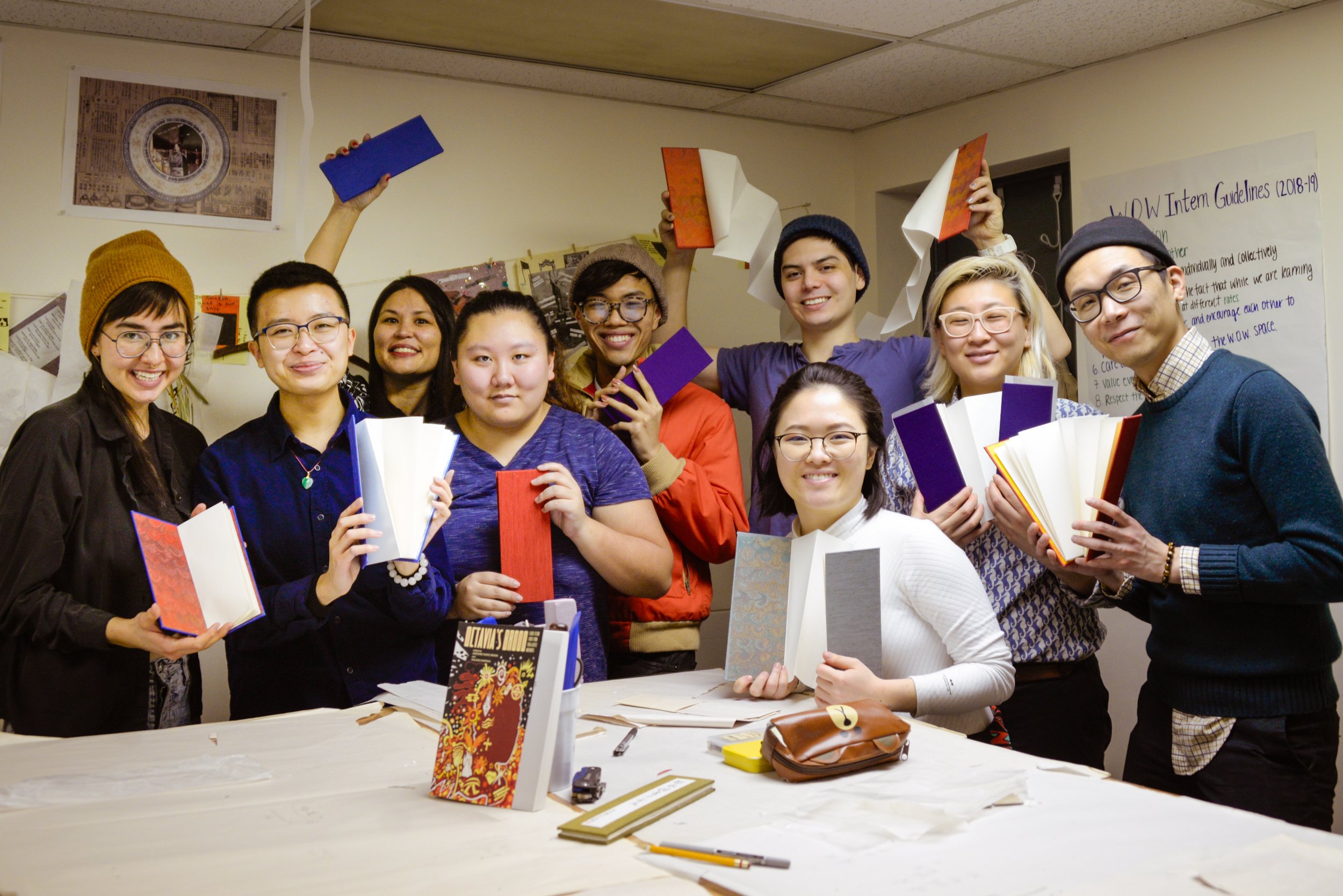 Group of diverse people holding open notebooks, smiling, standing in a room with posters and a whiteboard.