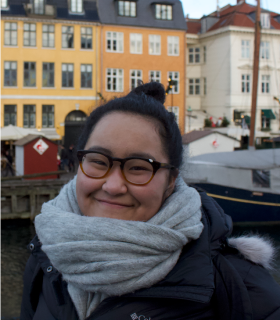 Smiling woman with glasses and a gray scarf near a harbor with colorful buildings and boats in the background.