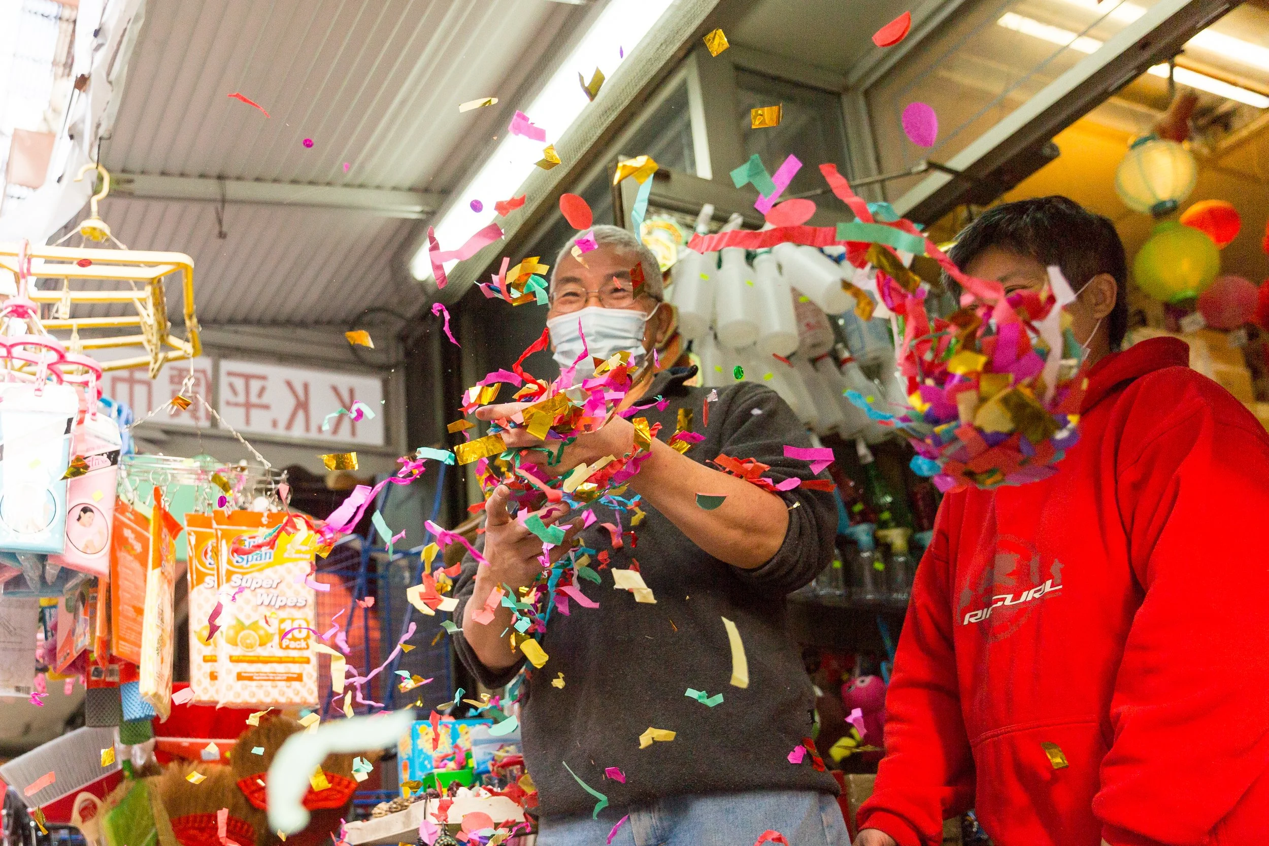 Two people at a store celebrating with colorful confetti. One person in a black jacket and face mask is holding a phone, and another in a red hoodie is standing nearby. Various items are displayed on shelves behind them.