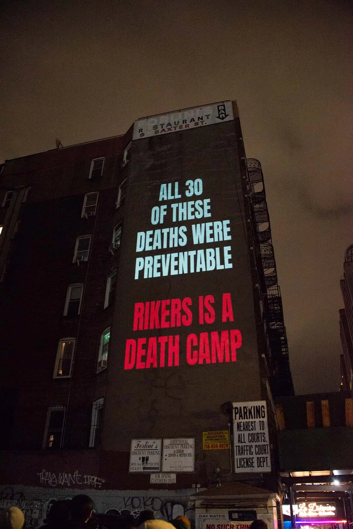 Large illuminated billboard on the side of a building displays a message about preventable deaths and bikers, with some smaller signs below. The message reads: "All 30 of these deaths were preventable. Bikers is a death camp."