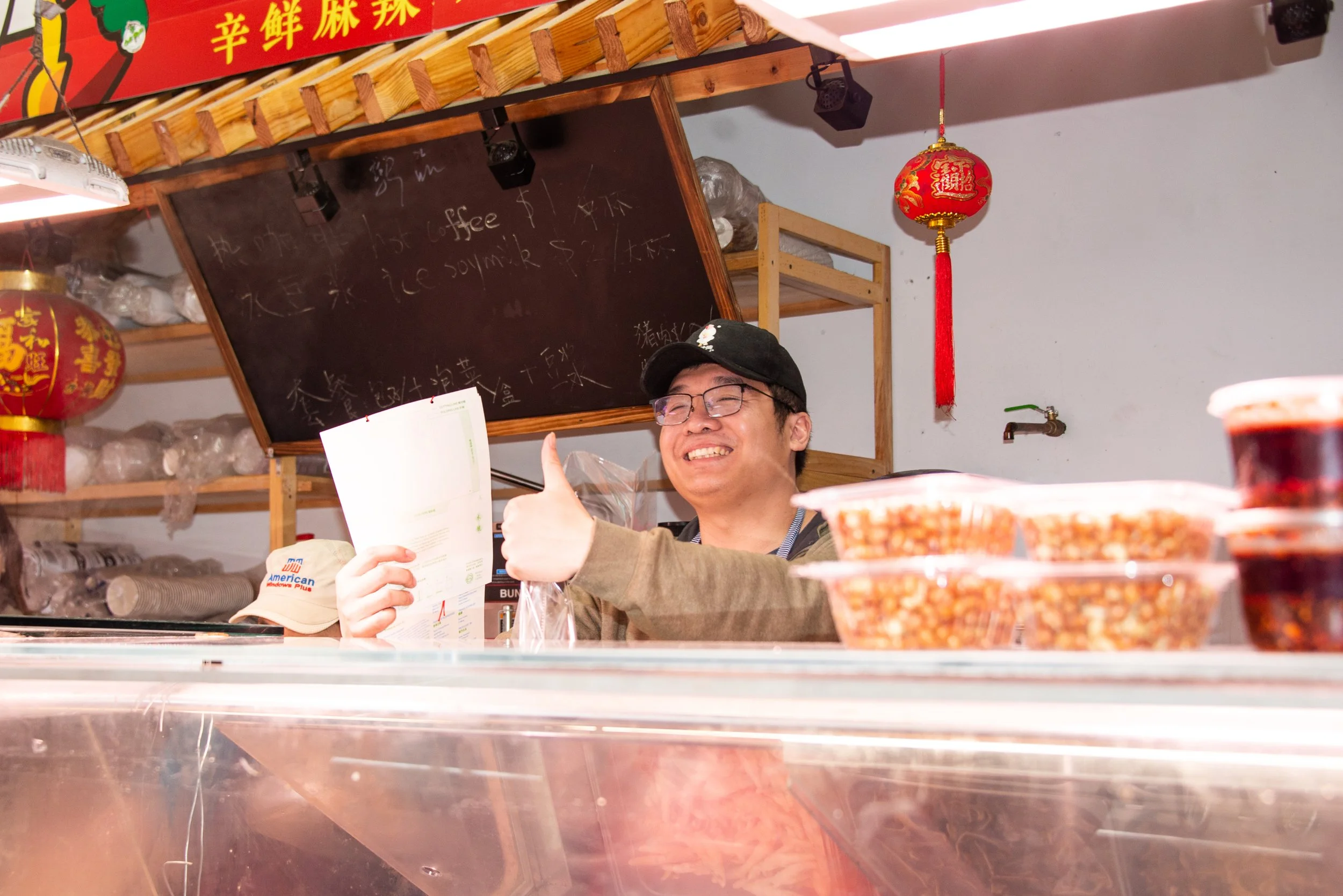 A cheerful man wearing glasses and a cap is giving a thumbs-up behind a food stall, with Chinese lanterns hanging to the side and bowls of food in front.
