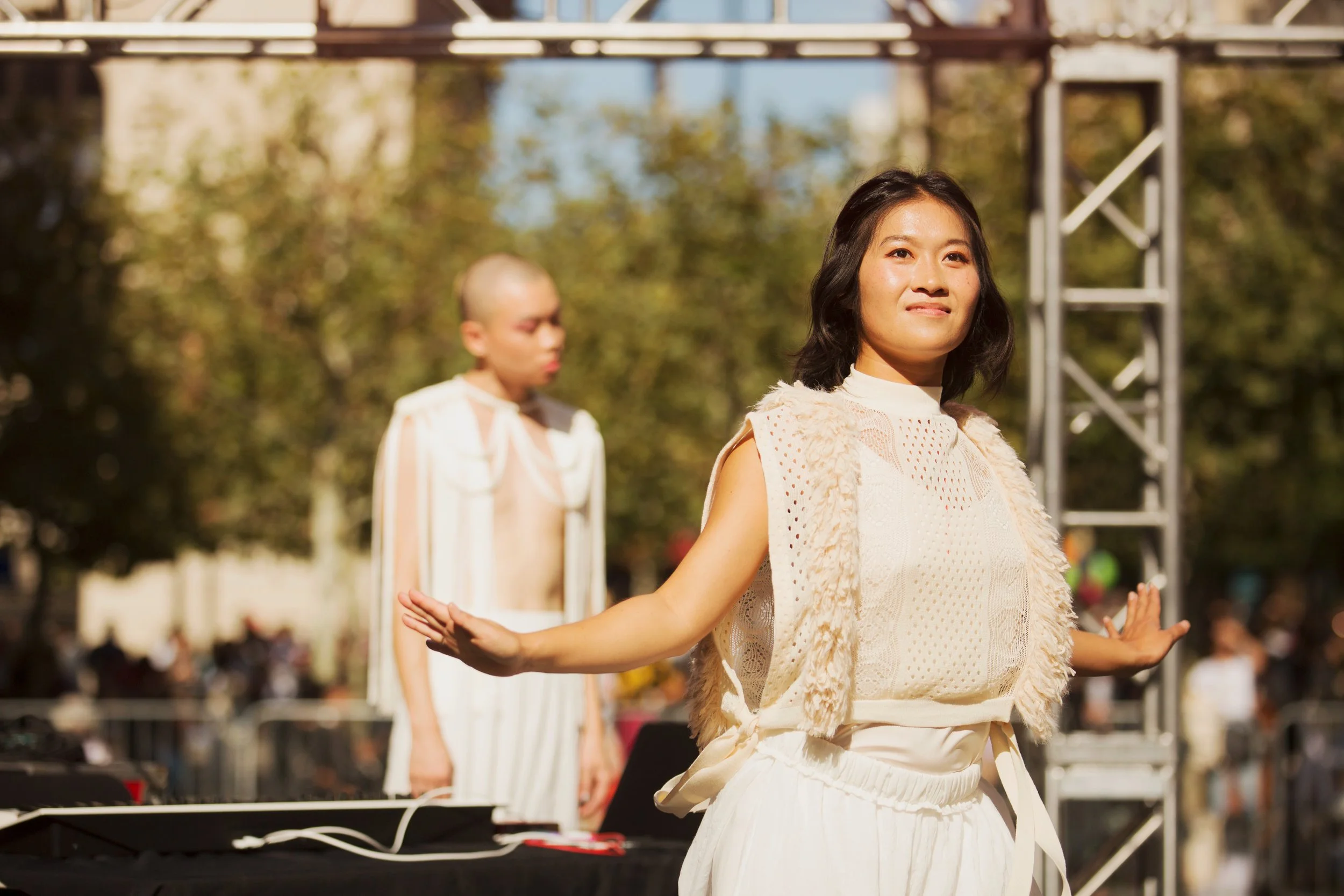 Two women, one with short dark hair and the other with a shaved head, performing outdoors on stage during the daytime. The woman in the foreground is smiling and has her arms outstretched, wearing a light-colored outfit with a textured vest. The woman in the background is focused, wearing a striped outfit, and standing near DJ equipment.
