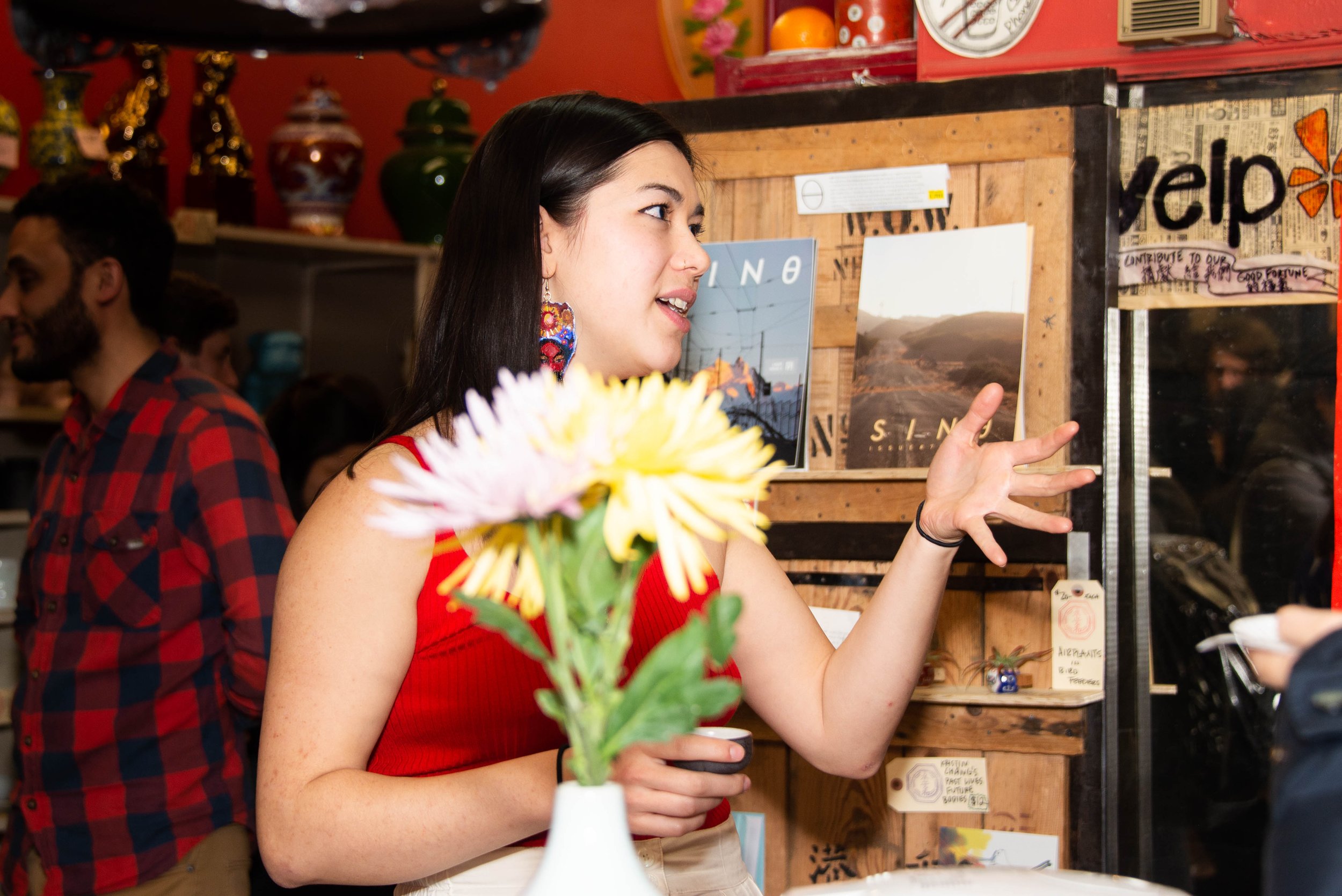 A woman in a red top talking and gesturing with her hand inside a colorful store, with vases and posters on the wall, and a bouquet of flowers in the foreground.