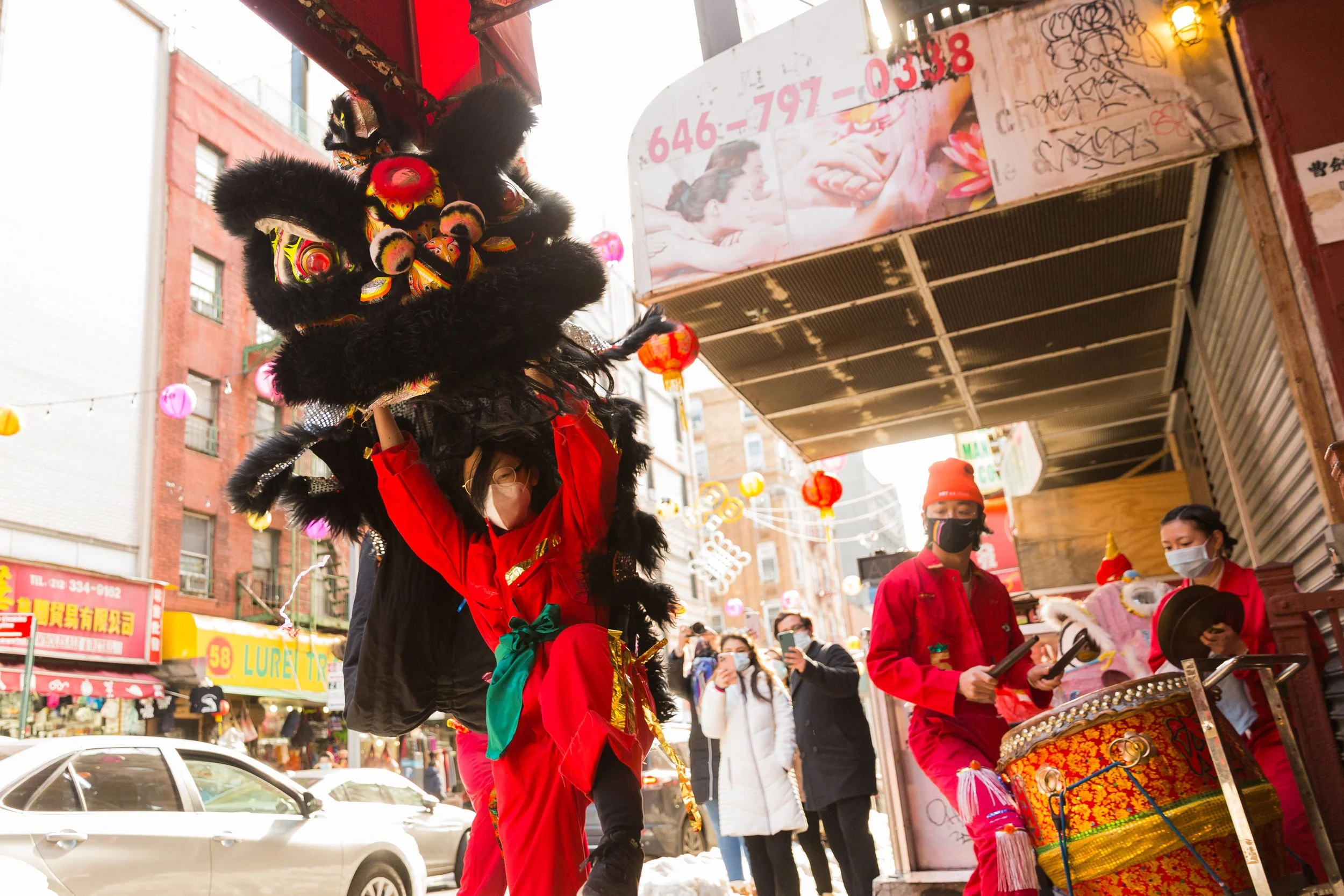 People celebrating a cultural event on a city street during daytime, with one person wearing a traditional lion dance costume, others playing drums, and onlookers taking photos, colorful lanterns hanging overhead.