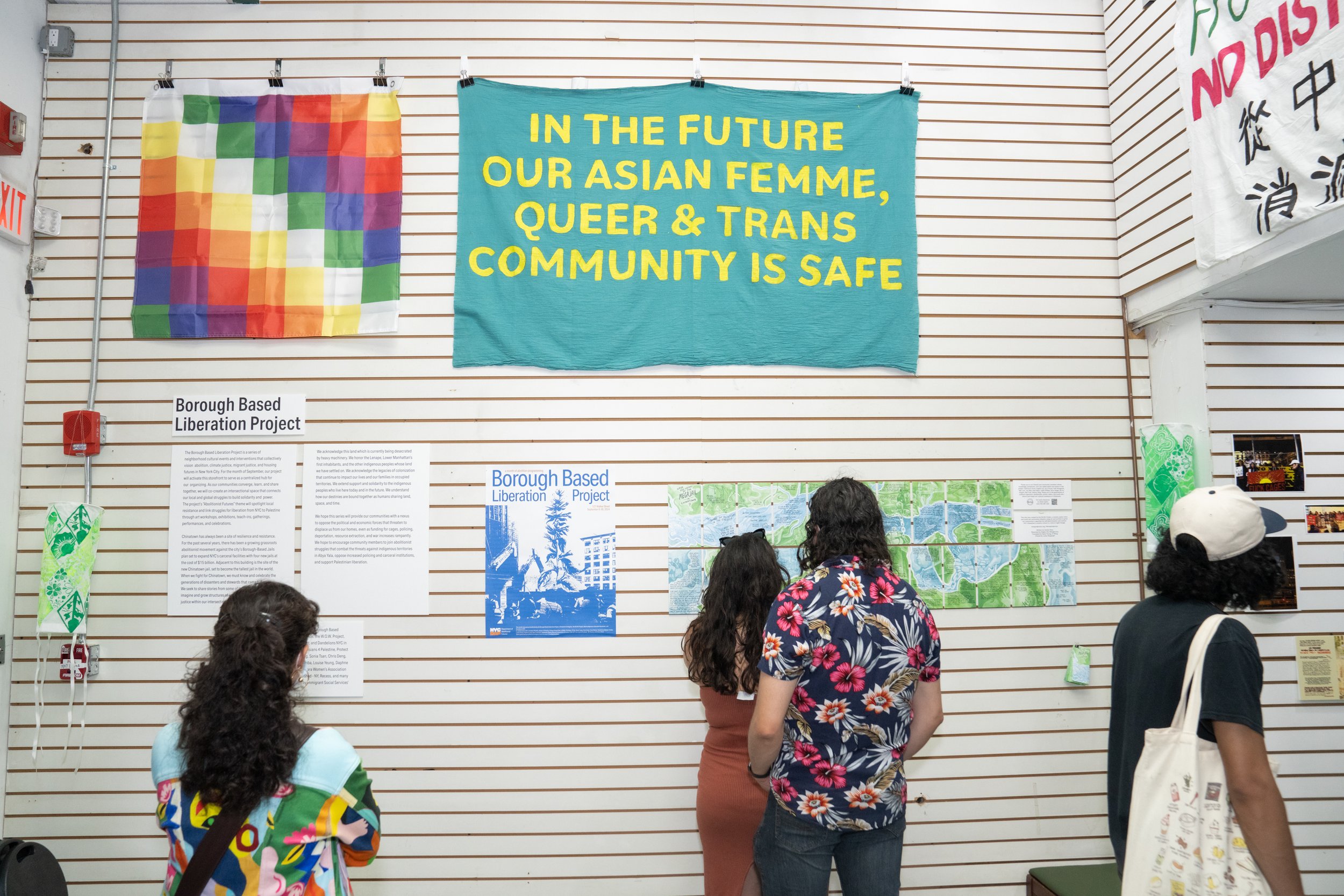 Four people standing and looking at display panels and posters in an exhibition. The background features colorful banners, including a pride flag and a large teal banner with yellow text that reads, 'In the future, our Asian femme, queer & trans community is safe.'
