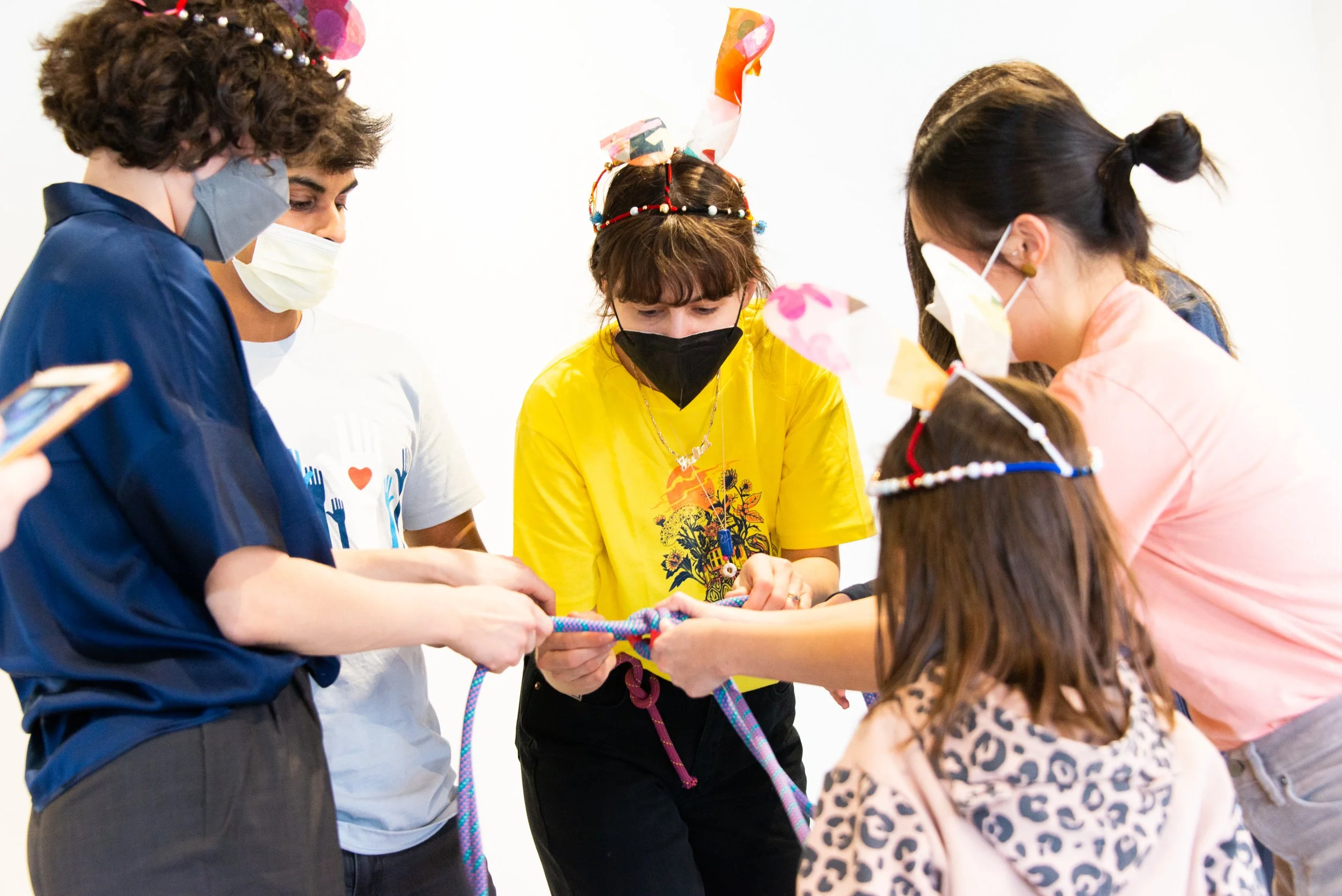 Group of children and teenagers wearing masks, gathered around holding a colorful rope, possibly playing a game or participating in an activity, with one girl wearing a decorated headband with bunny ears and a yellow shirt.
