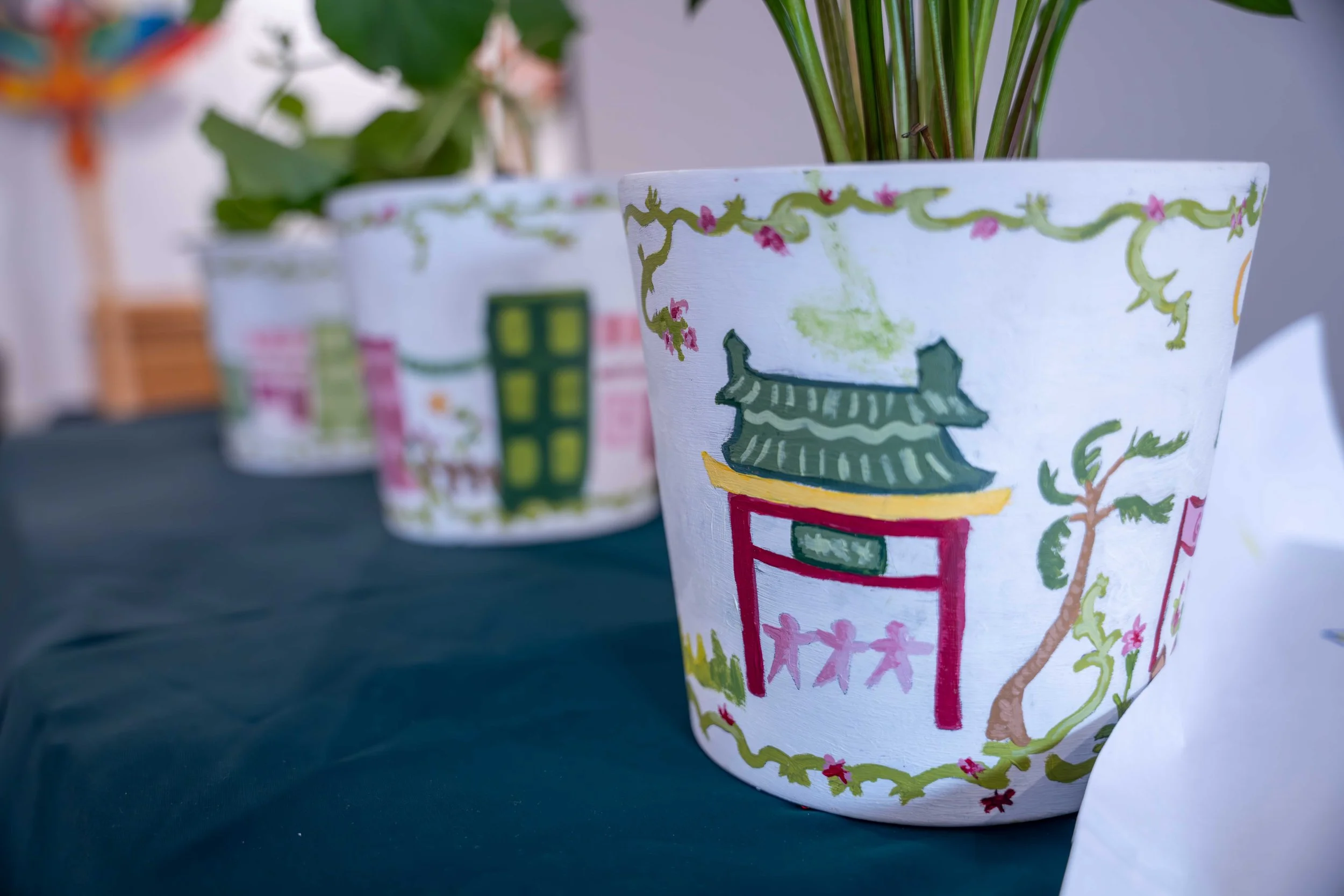 Hand-painted flower pot with a pagoda-style building, trees, and decorative borders, placed on a table with other similar painted pots in the background.