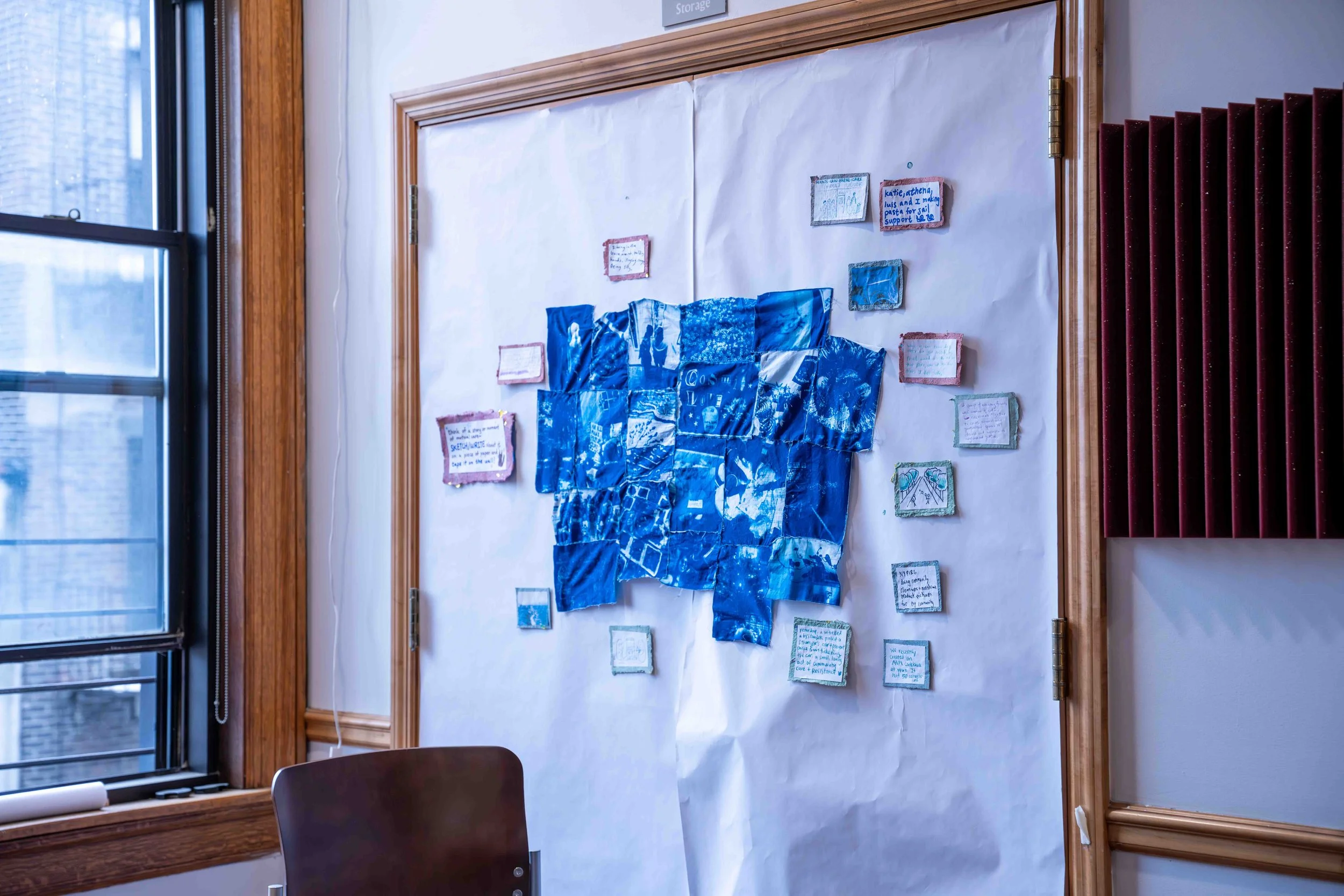 Display on a wall with blue fabric arranged in a collage, surrounded by small framed notes and artwork, in a room with wooden trim, a large window, and acoustic panels on the wall.