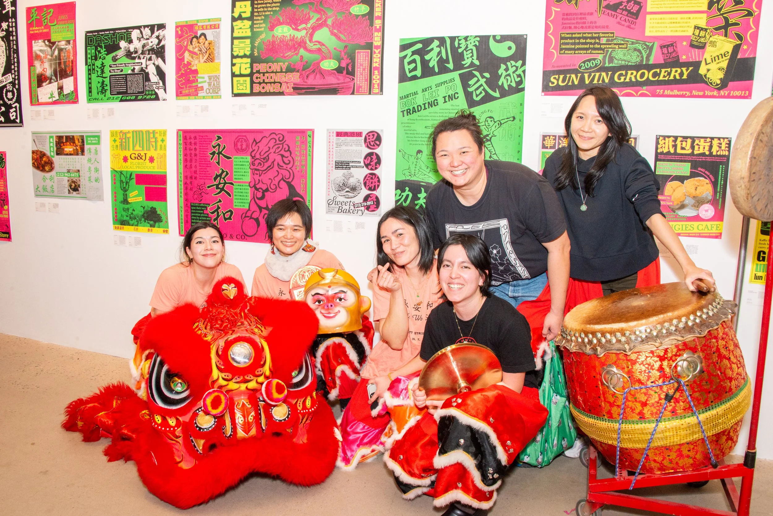 Group of six women posing with a traditional Chinese lion dance costume, drum, and cymbals in front of colorful posters on a gallery wall, celebrating Chinese culture.