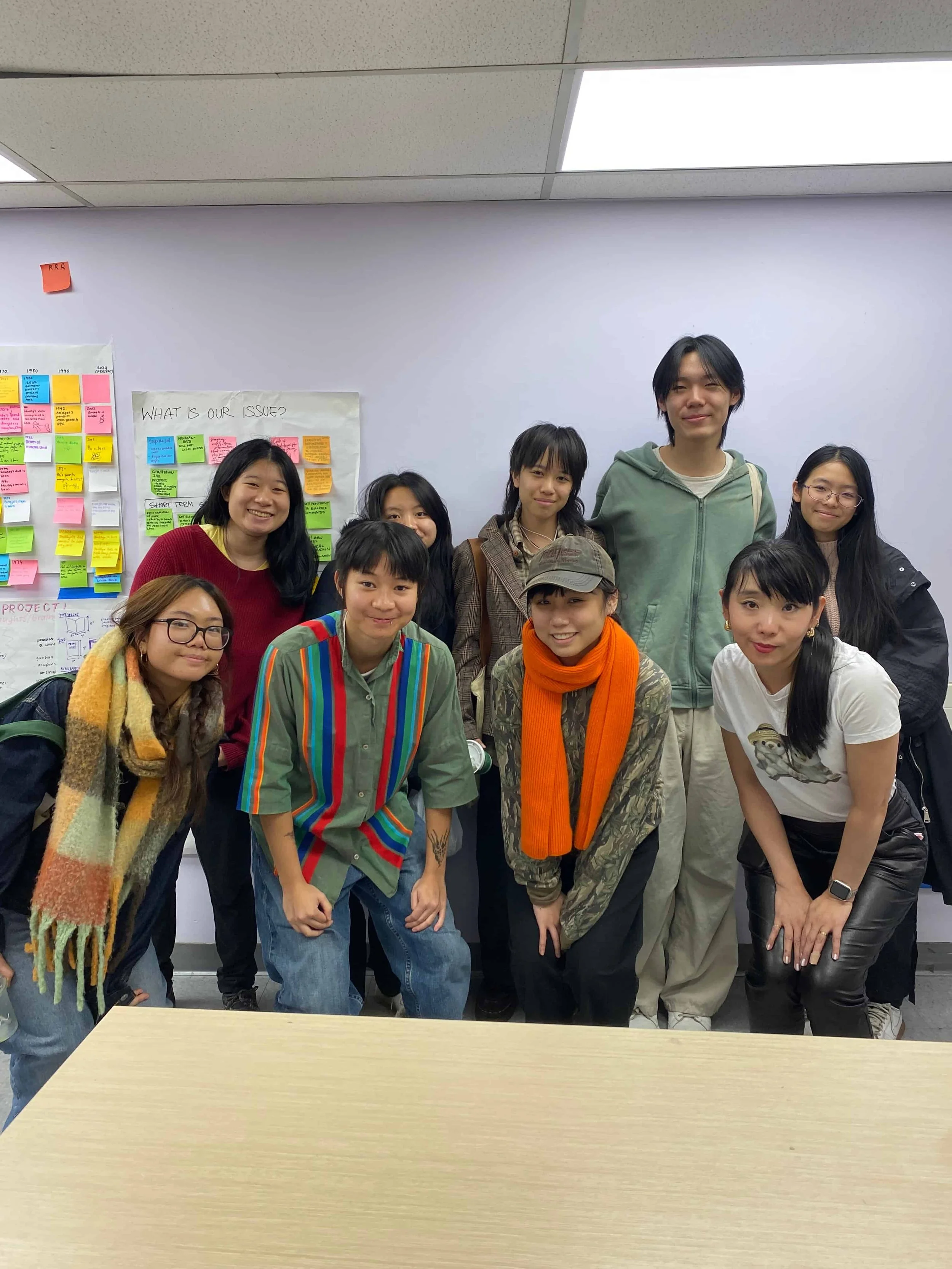 Group of ten young people standing in a classroom, smiling, with sticky notes and charts on the wall behind them.