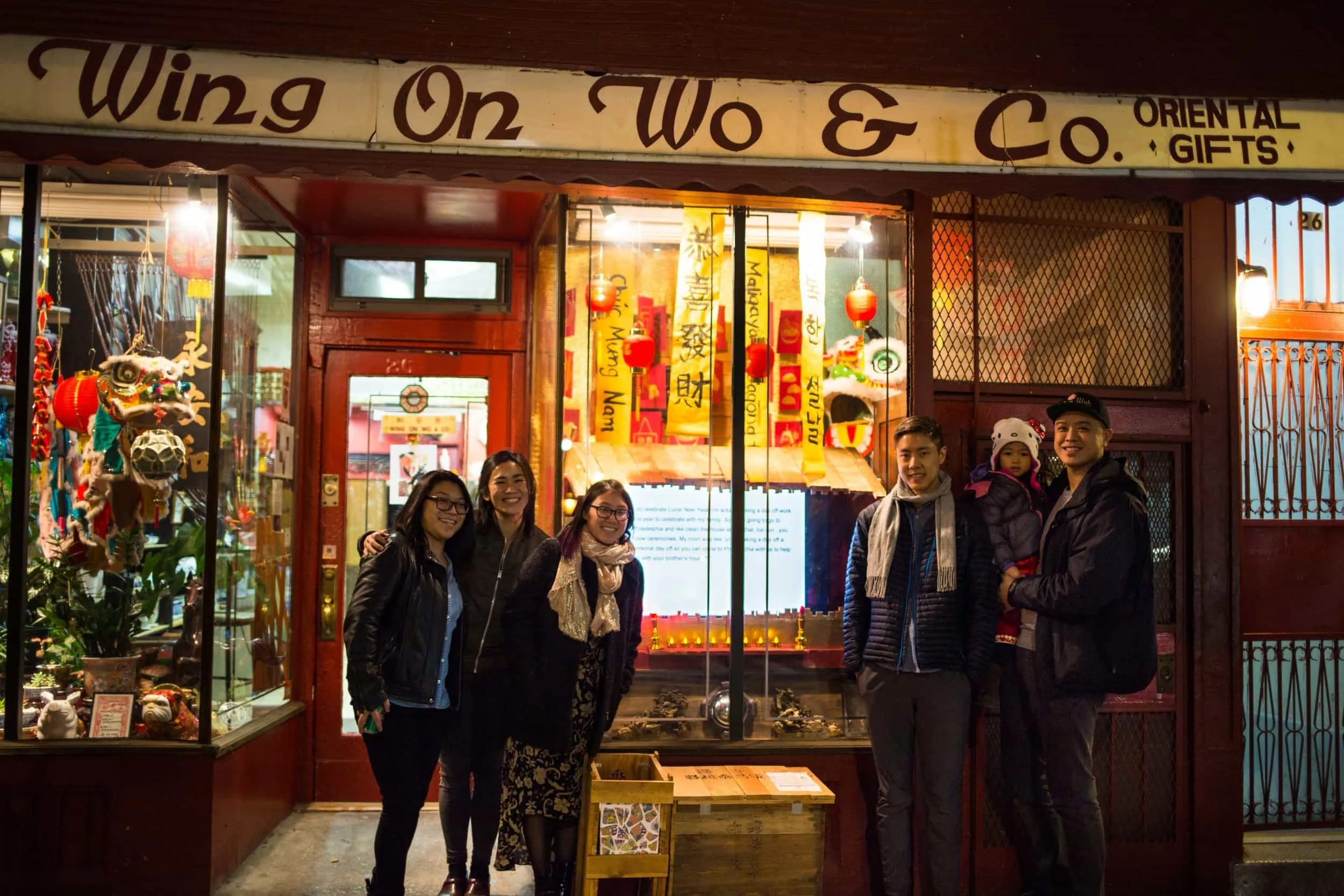 A group of six people, four women and two men, standing outside a shop named Wing On W & Co. Oriental Gifts, smiling at the camera. One man is holding a young girl in a pink and purple jacket and white hat. The shop window displays Japanese and Chinese decorations, lanterns, and souvenirs.