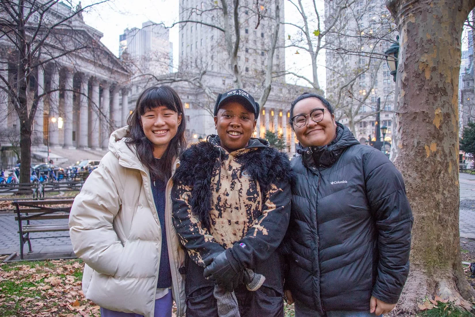Three smiling women standing outdoors near a tree with city buildings in the background.