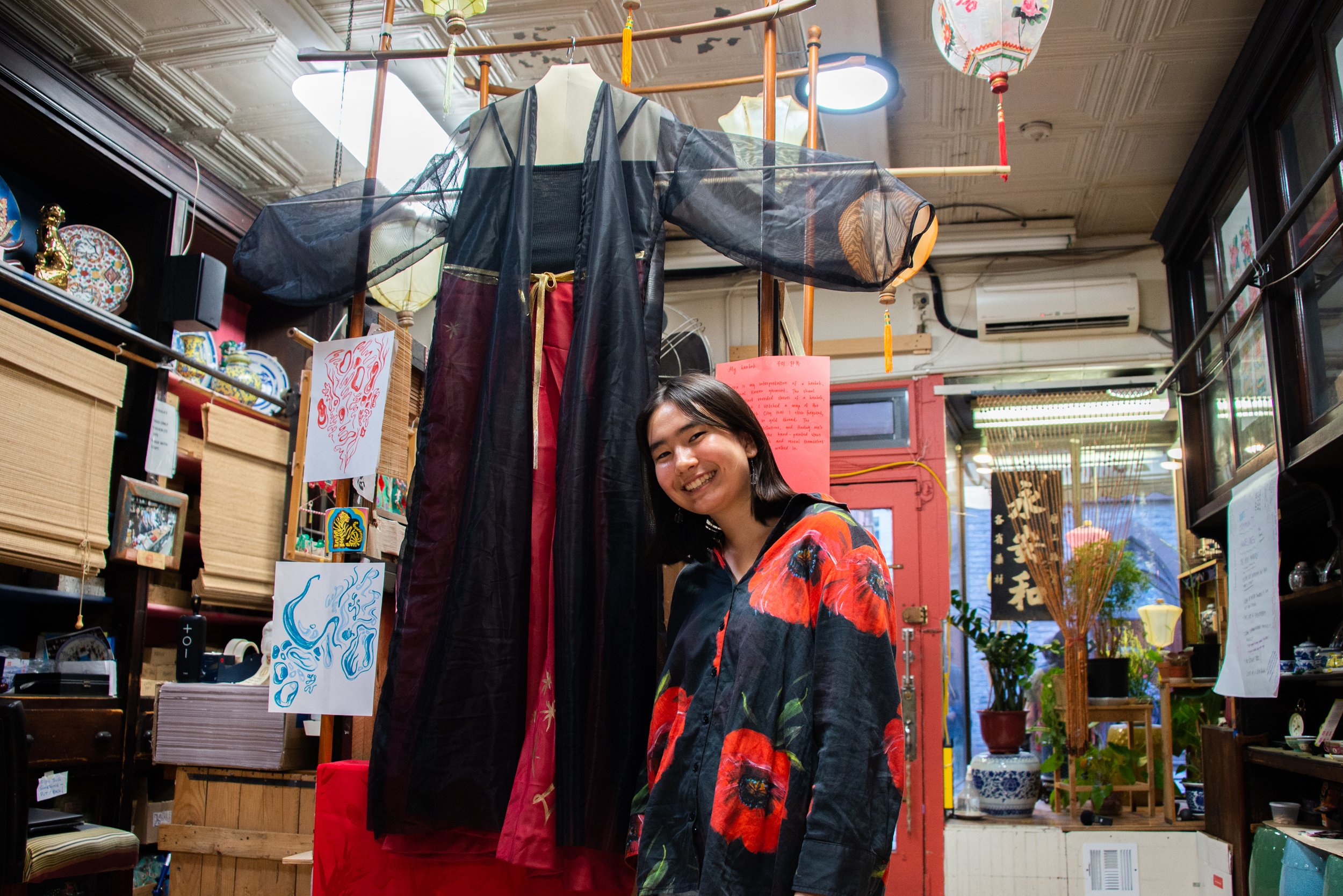 A young woman standing inside a colorful shop with traditional Japanese clothing and decorations, smiling at the camera. There is a large kimono hanging behind her, artwork on display, and various decorative items and plants around the space.