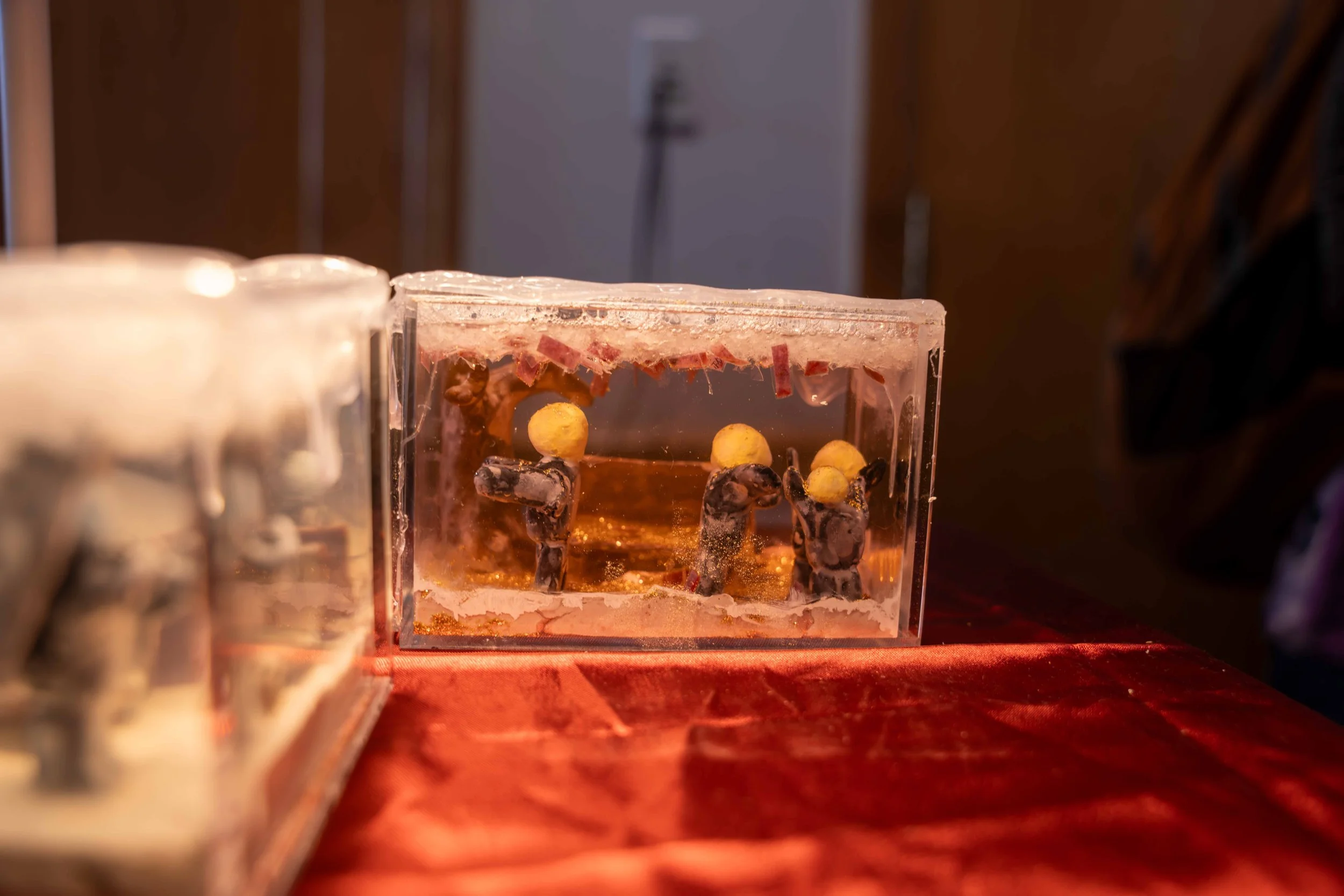 Close-up of a Christmas ornament featuring three miniature figures of monkeys hanging from a tree branch, inside a transparent box with a wooden surface underneath.