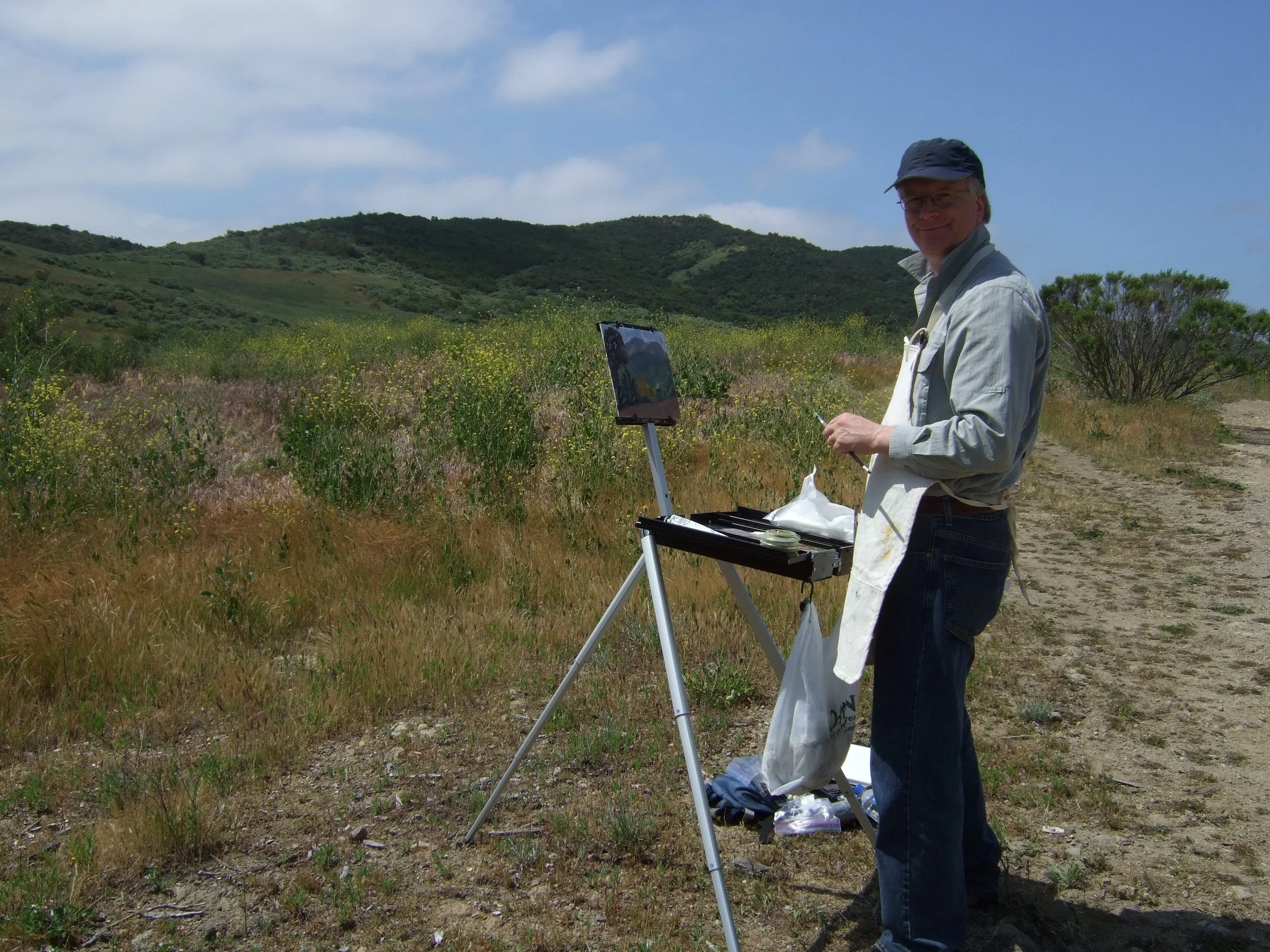 Dan Loyd painting outdoors in a scenic grassy landscape with hills in the background.