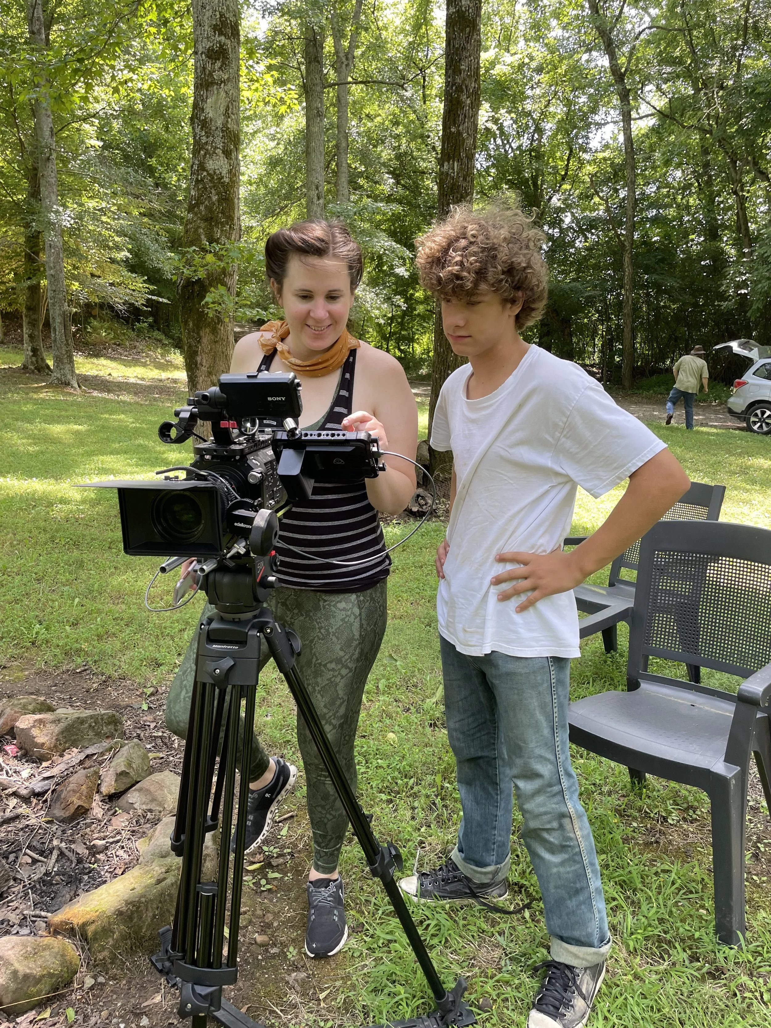 Two people outdoors with video camera on tripod, trees in background.