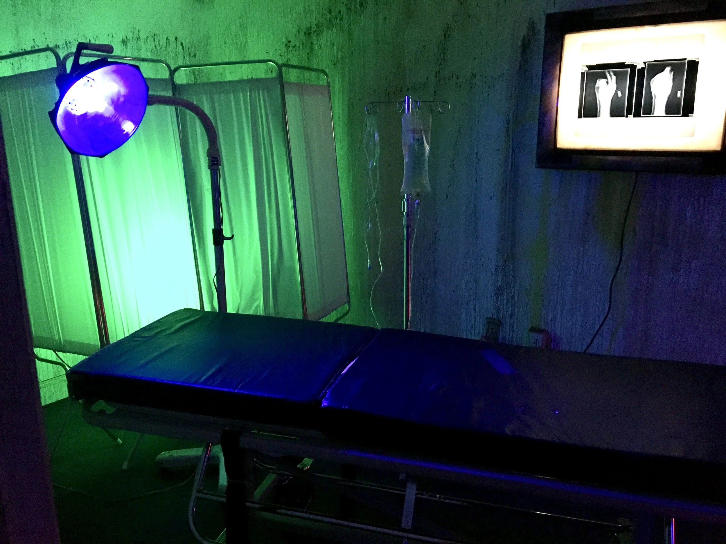 Dark medical room with an examination table, surgical light, IV stand, and illuminated X-ray display.