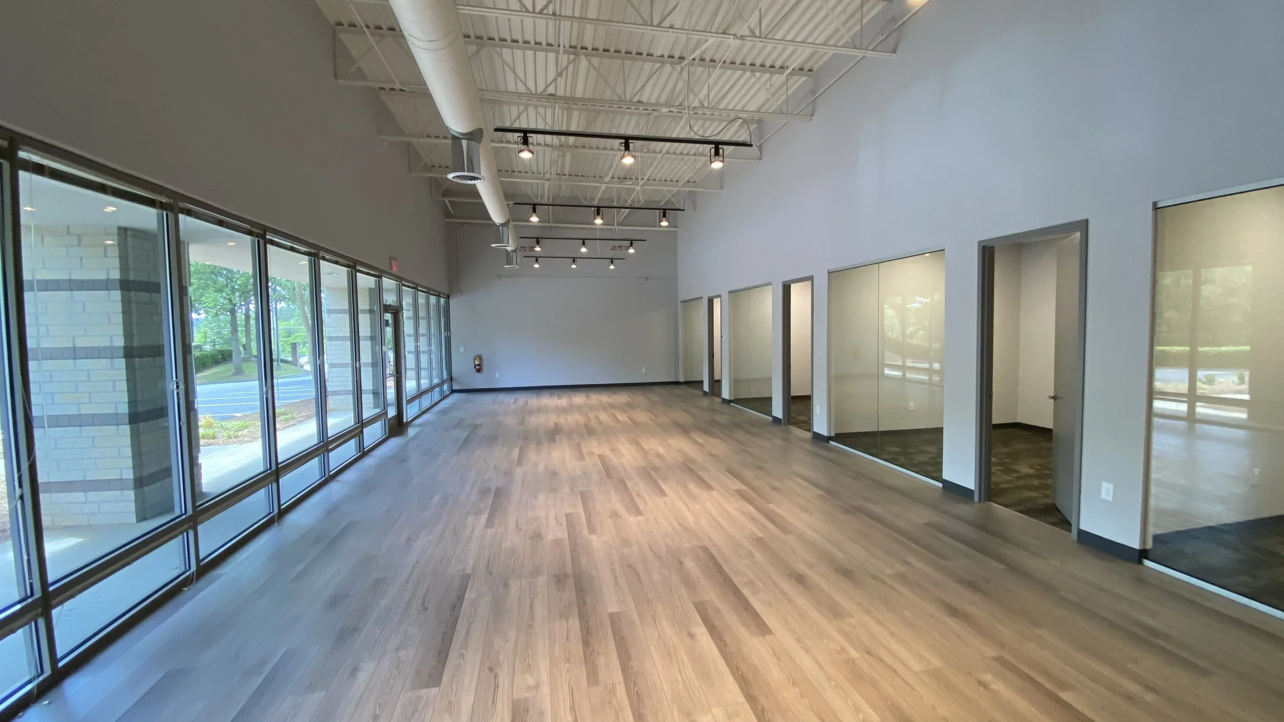 Empty commercial space with hardwood floors, large windows, and multiple glass-door offices on one side.
