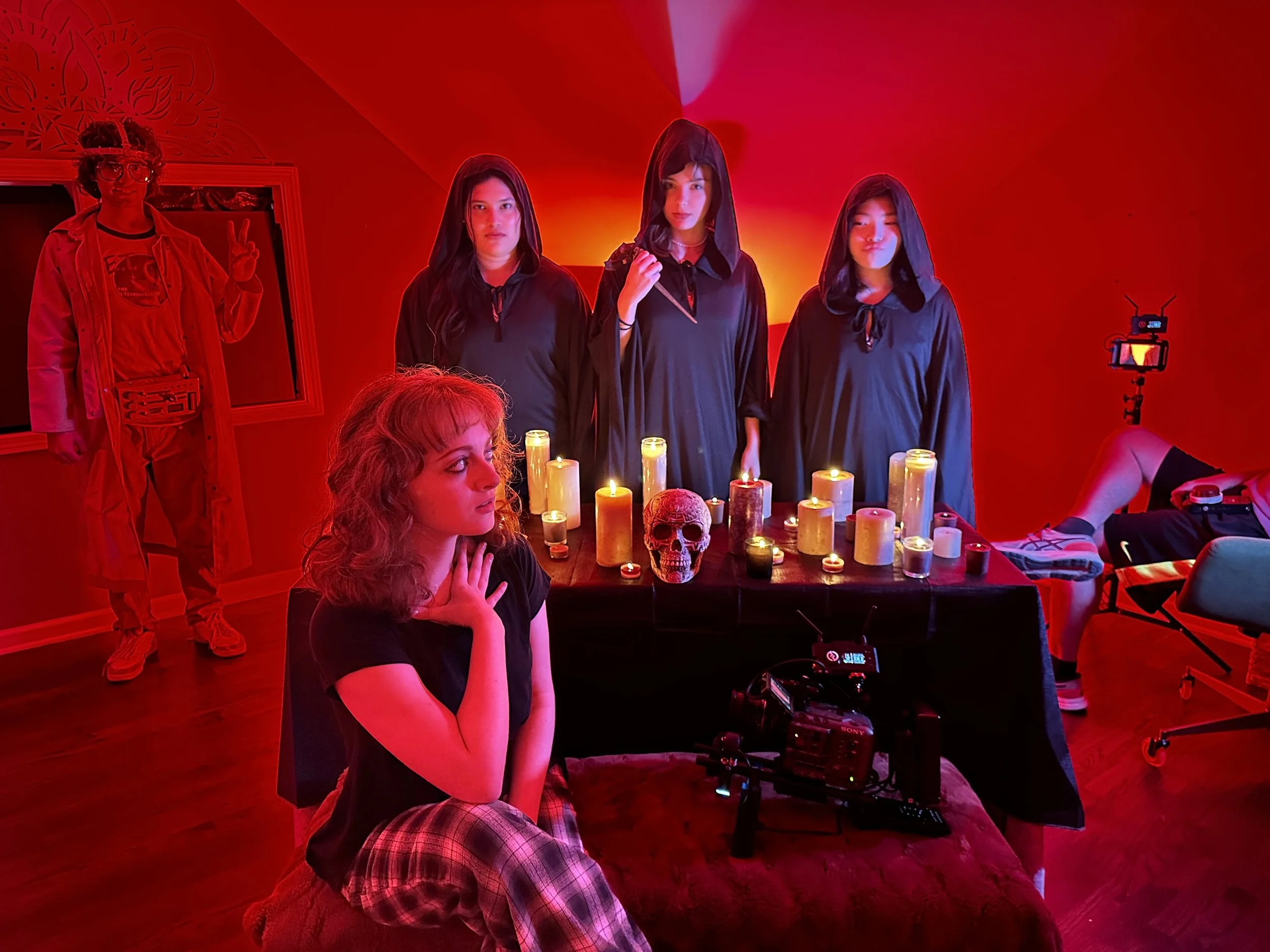 Group of people in a dimly lit room with red lighting, dressed in dark robes, surrounding a table with candles and a skull. A person sits in the foreground with a thoughtful expression. A stand with a mounted camera is visible, suggesting a film or photography setup.