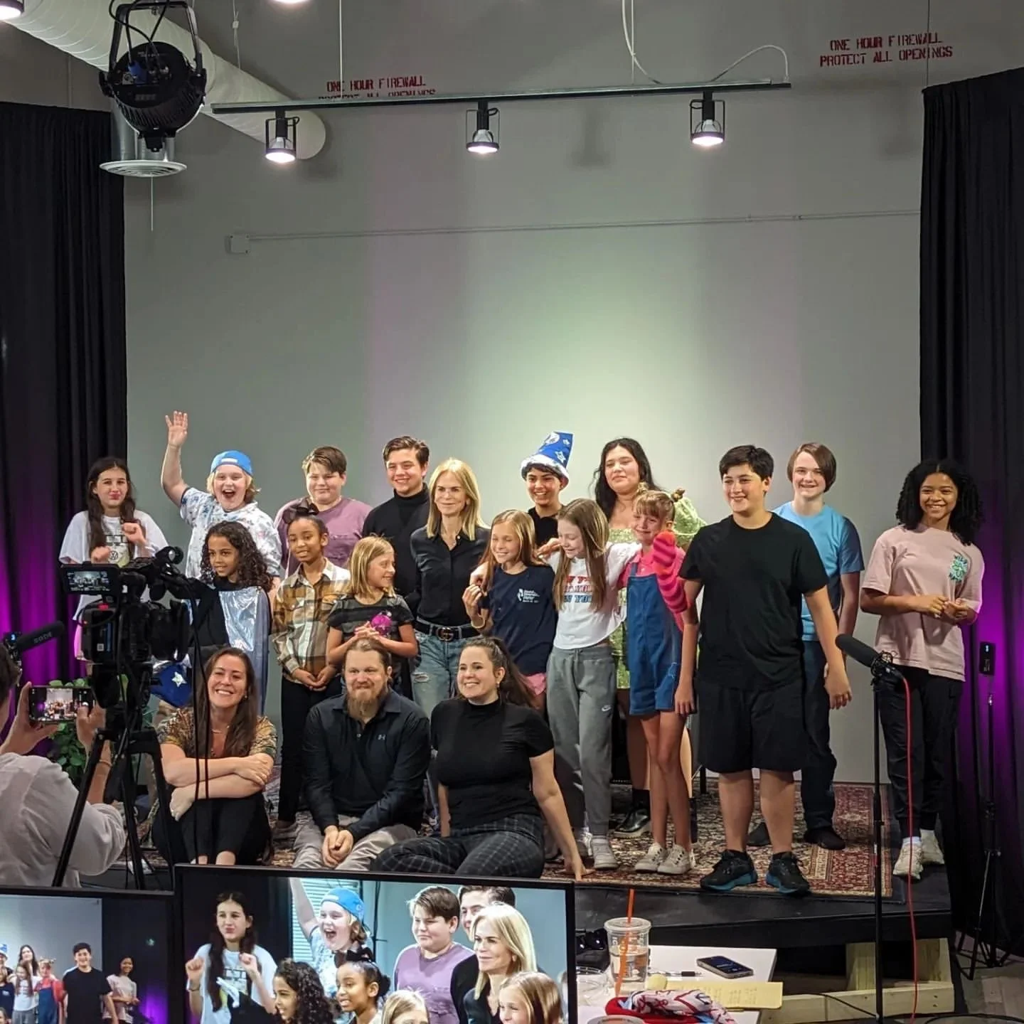 A group of people, including children and adults, pose for a photo on a stage in a studio setting. Some are standing and others are sitting. A camera is visible, suggesting a video or photo shoot.