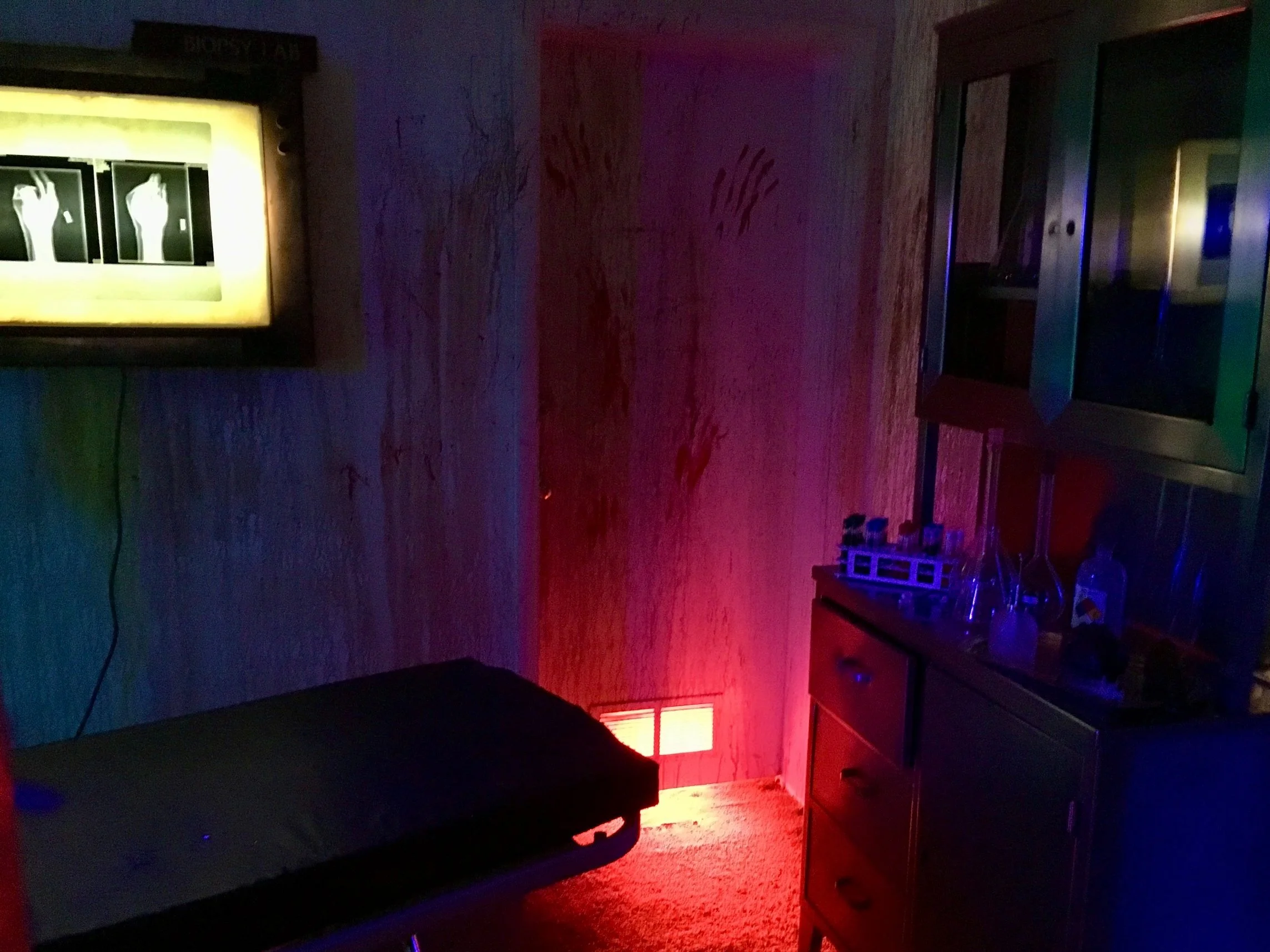 Dark, eerie laboratory room with x-ray lightbox, examination table, and cabinets with scientific equipment.