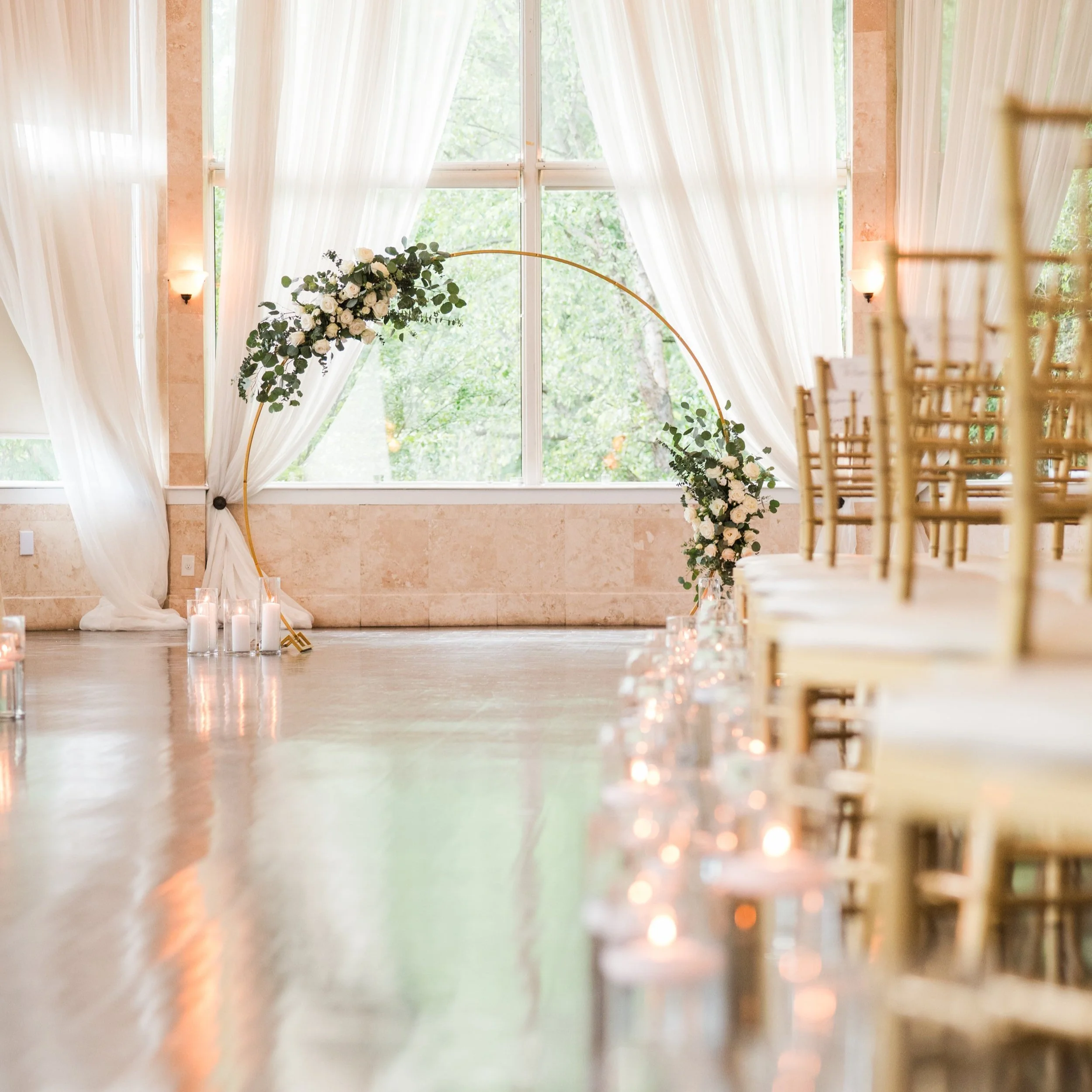 Indoor wedding ceremony setup with a circular floral arch, gold chairs, draped white curtains, and lit candles along the aisle.