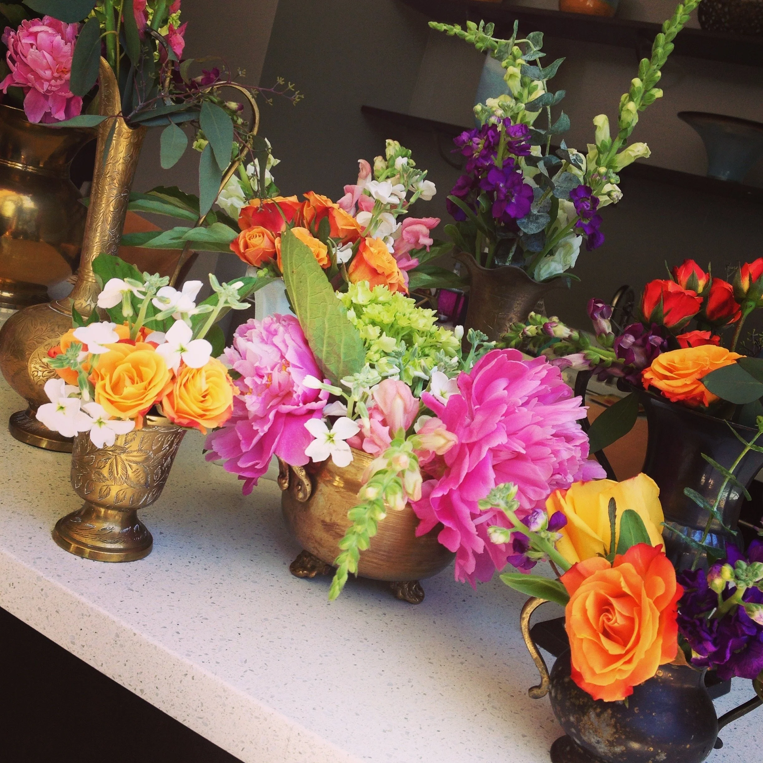 Assorted colorful flowers in decorative brass vases on a countertop.