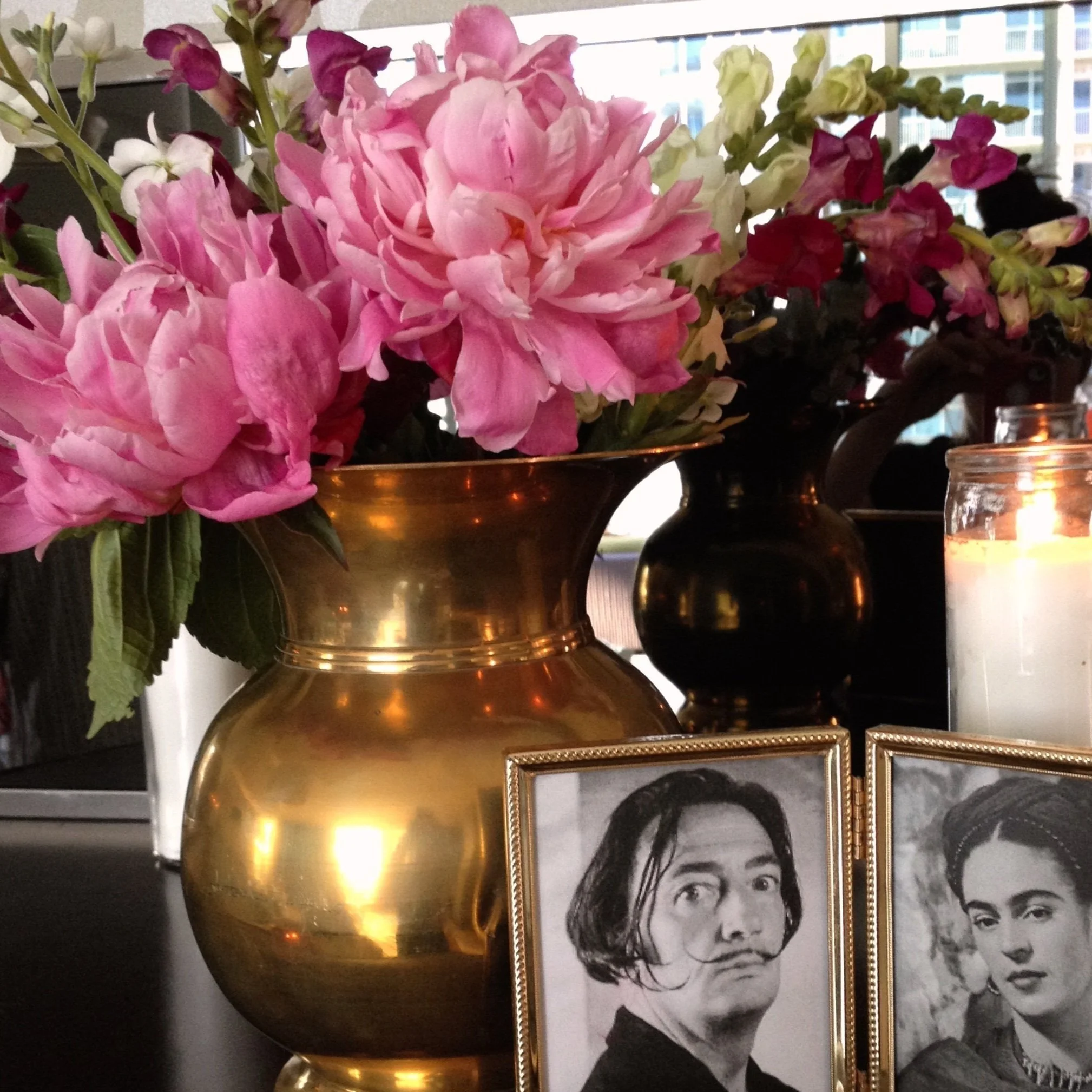 A brass vase with pink and white flowers on a table, next to black and white framed portraits of two famous artists and a lit candle in a glass holder.