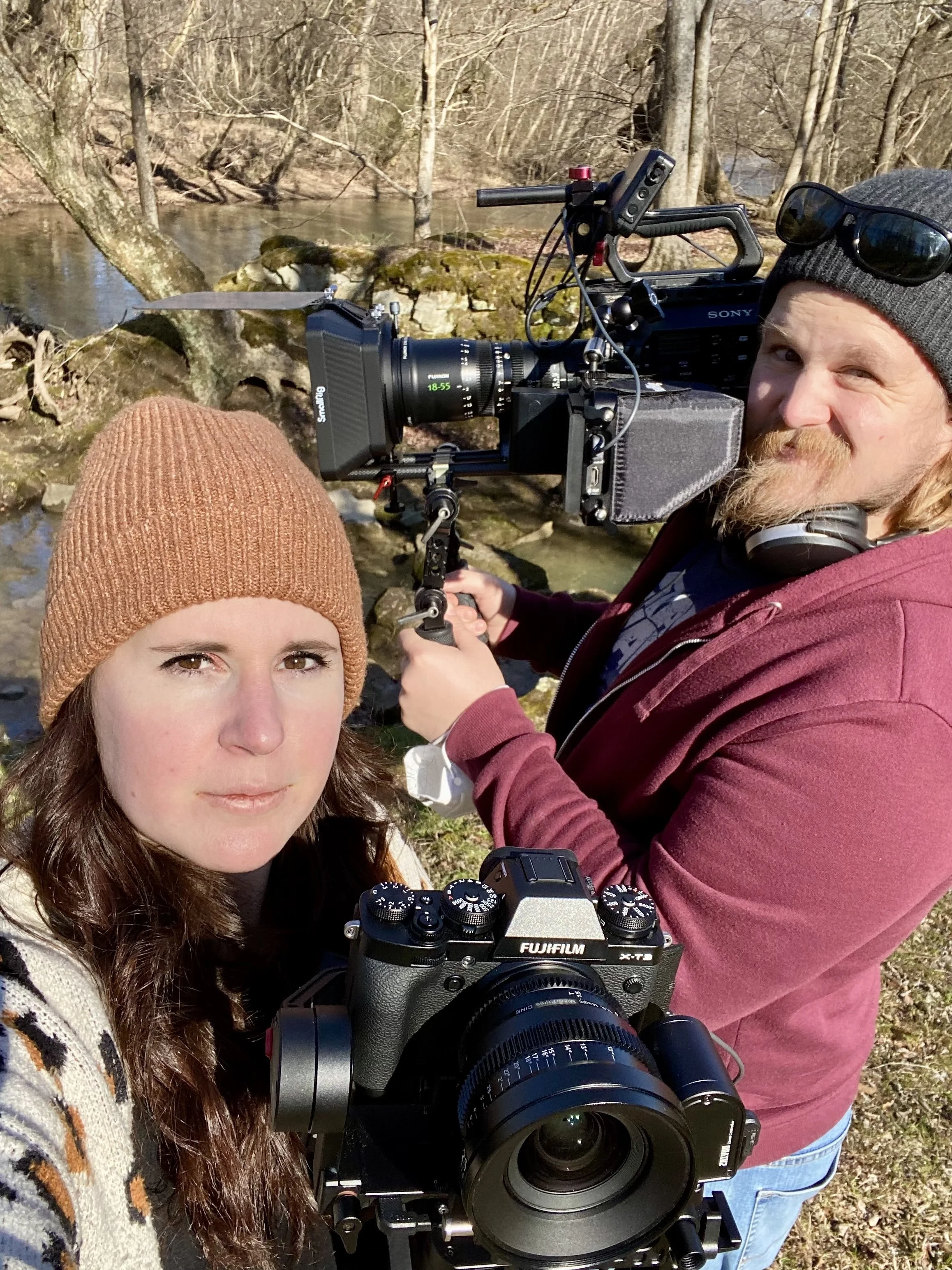 Two people outdoors with camera equipment near a stream, surrounded by bare trees. One holds a large Sony camera, the other a Fujifilm camera. Both wearing knit hats.