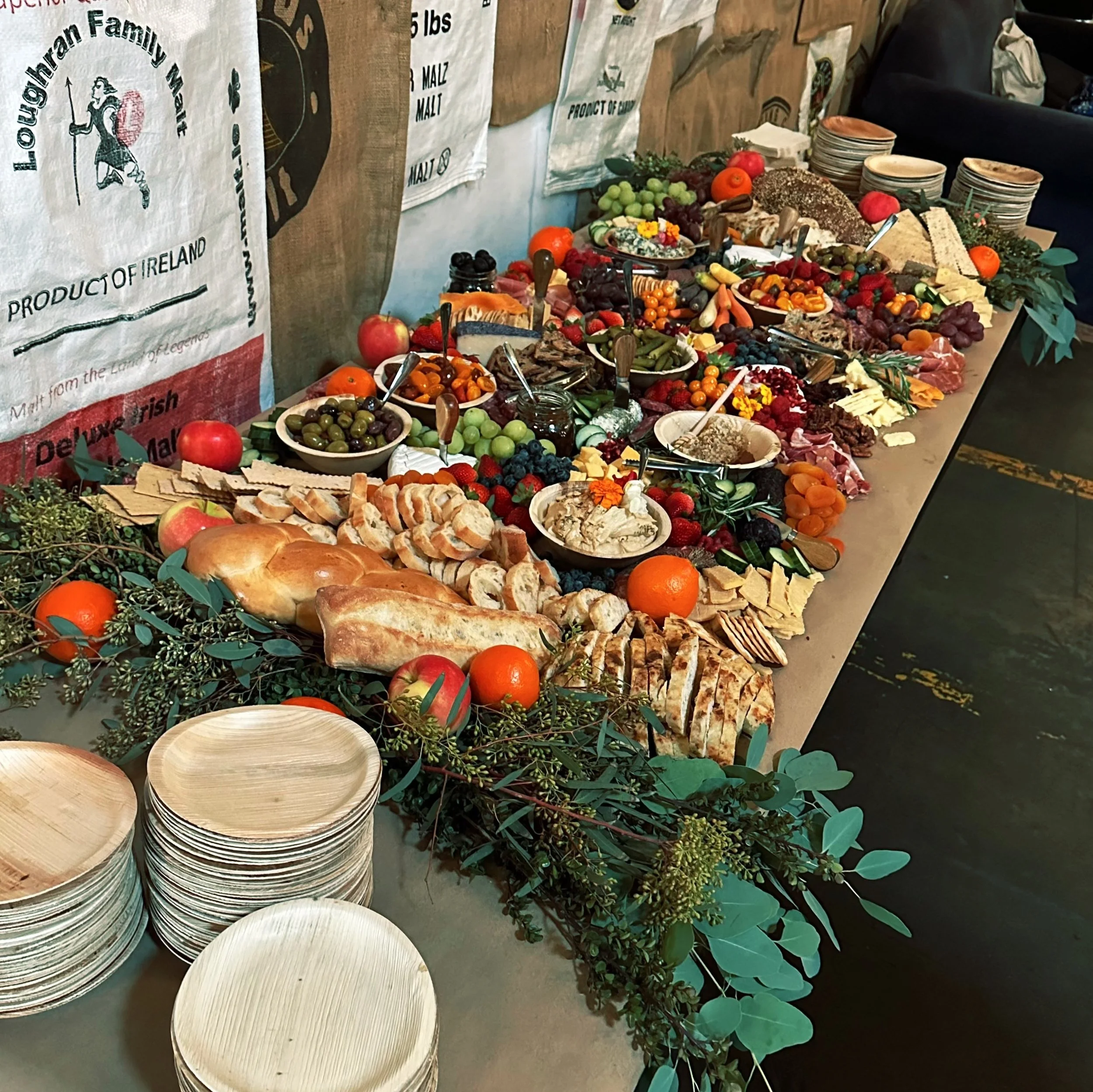 A wooden table set with a variety of foods, including sliced bread, various cheeses, fruits like grapes and oranges, olives, nuts, and a selection of meats. Plates are stacked at one end, and greenery is used for decoration. The background displays sacks with branding.