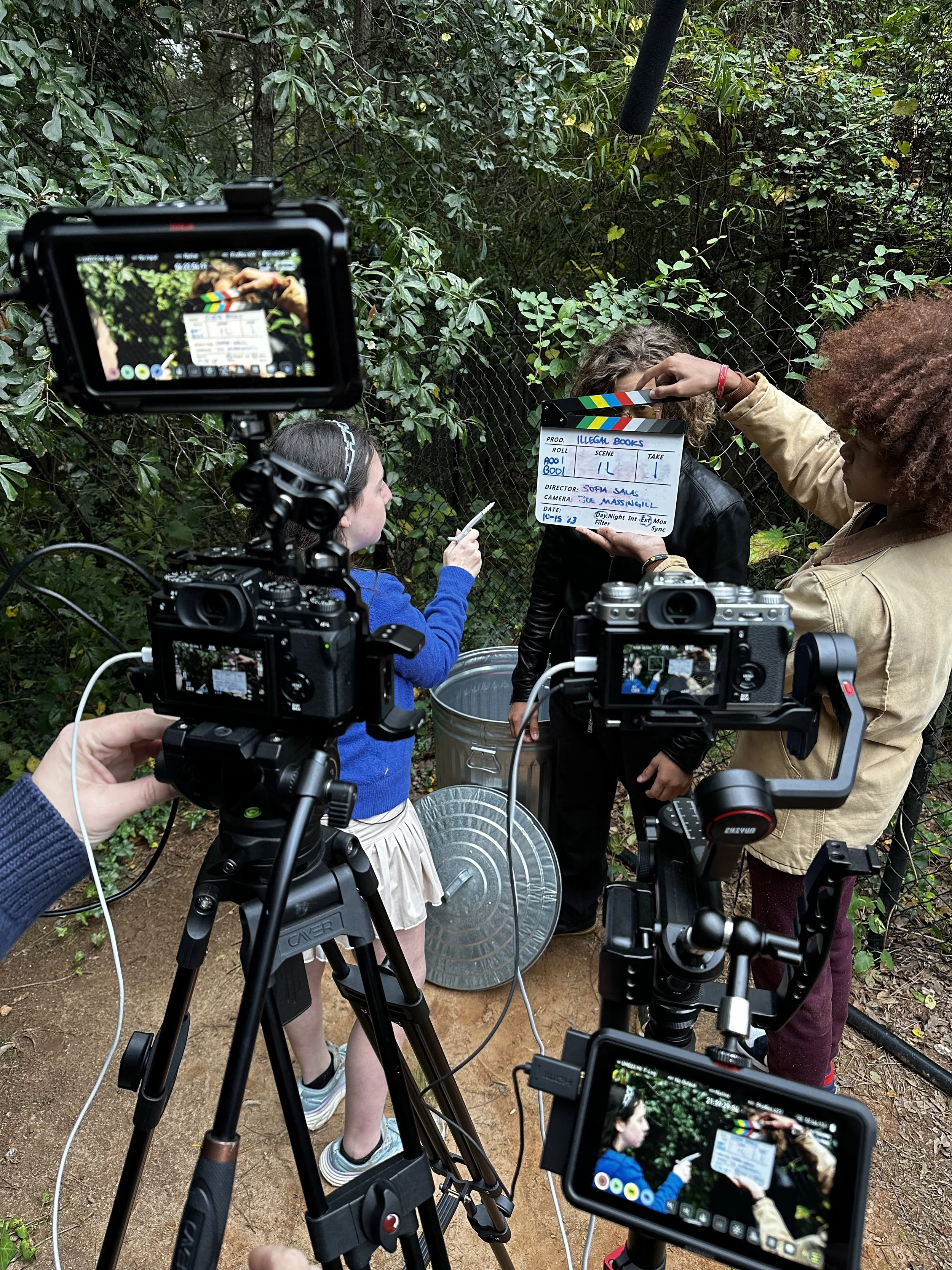 Behind-the-scenes film set with multiple cameras focusing on two actors and a clapperboard in an outdoor setting.