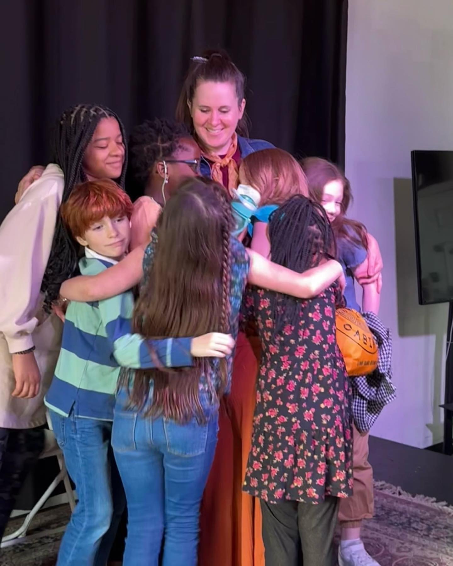 Group hug by several children and an adult, all smiling, on stage after a show.