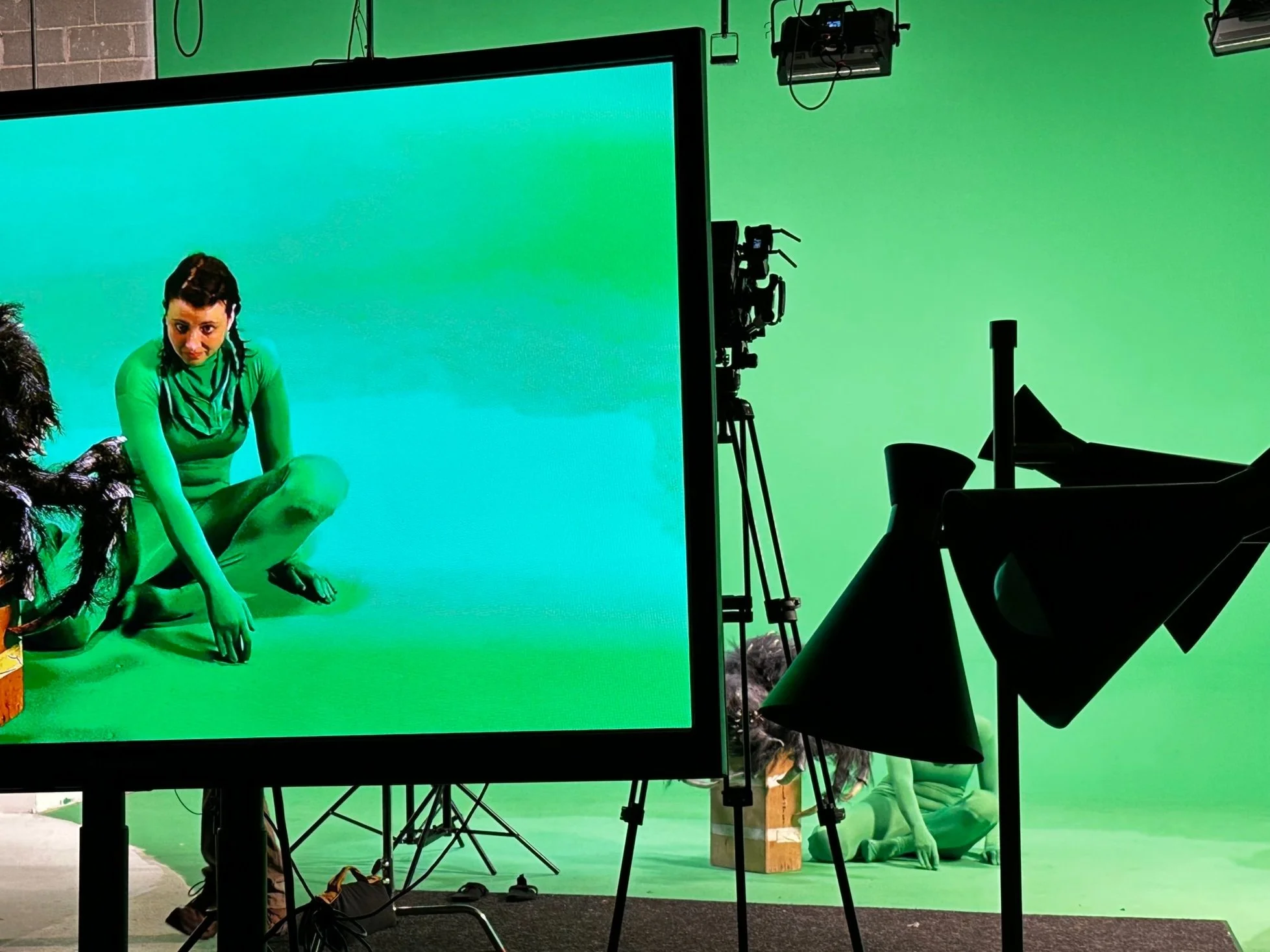 Person in green bodysuit on a film set with green screen background, surrounded by cameras and lighting equipment.